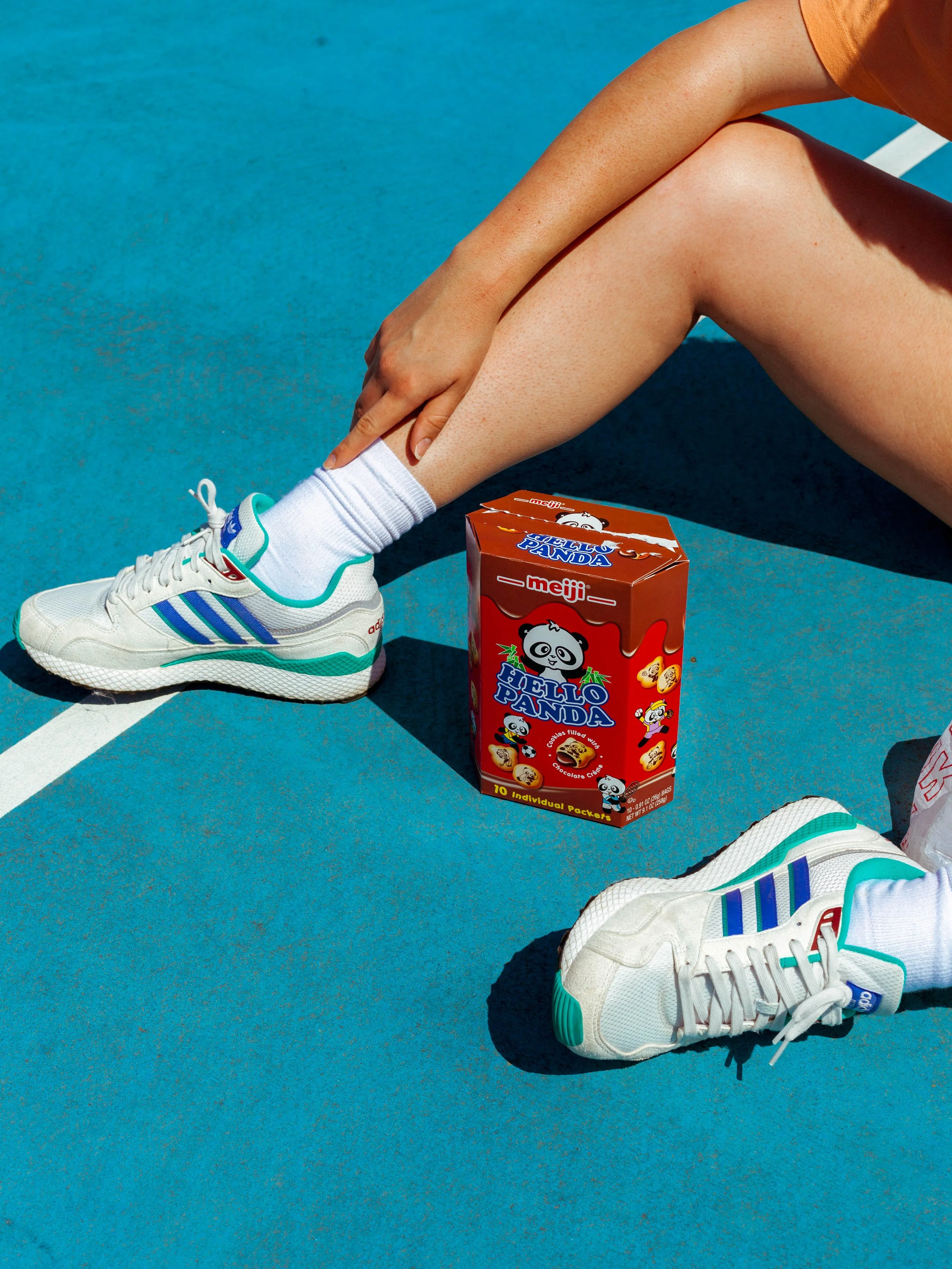Person in white sneakers with blue and green accents, white socks, and orange shirt sitting on a blue outdoor court, with a red boxed Hello Panda snack nearby.