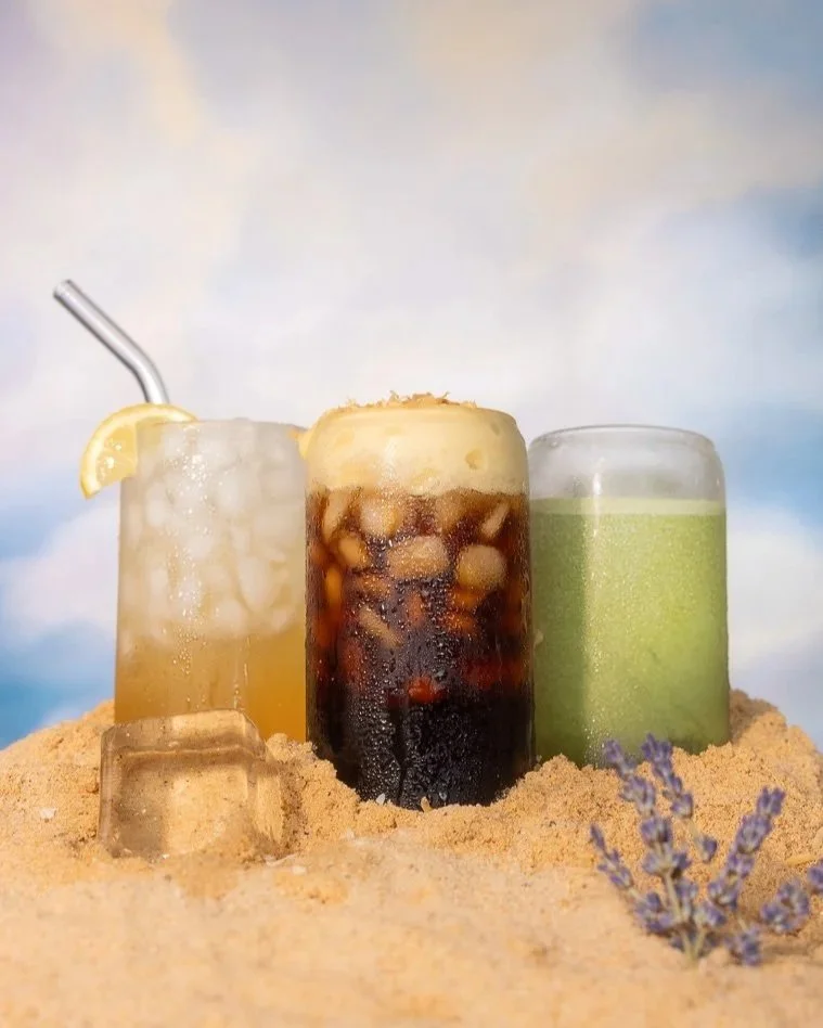 Three cold beverages in glasses on sandy ground with purple flower, ice cube, and pastel sky background.