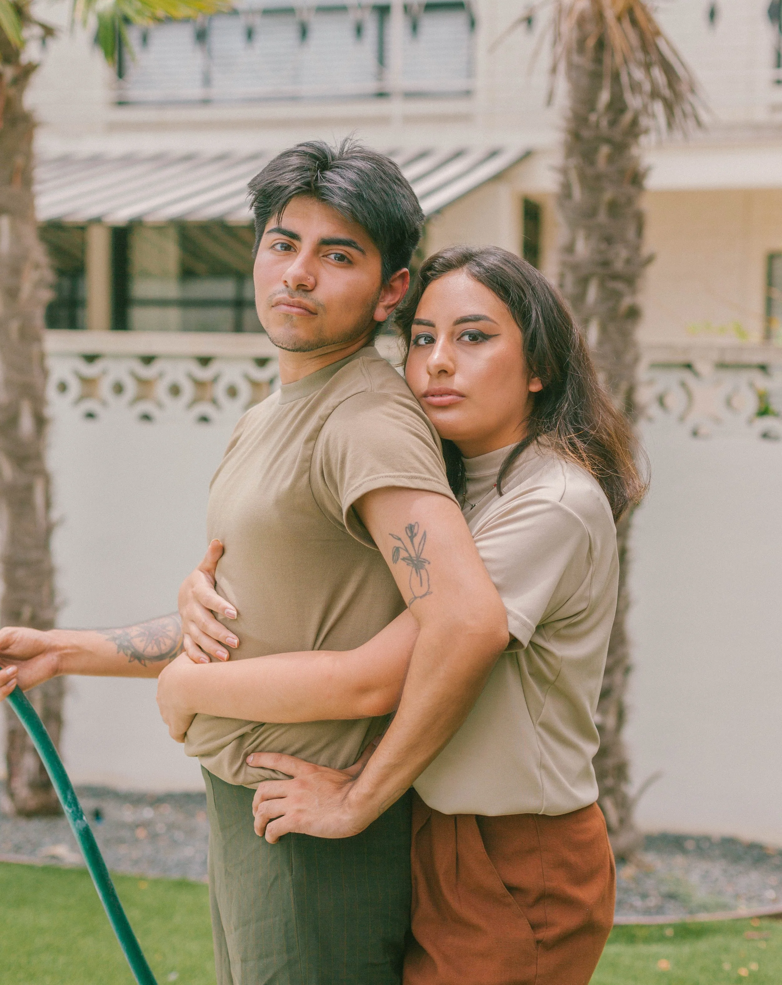 woman has her arm around the man's waist. The background shows a house with a balcony and some trees. The man has dark hair and is wearing a beige t-shirt, while the woman has dark wavy hair, makeup with winged eyeliner, and is wearing a light-colored shirt and brown pants.