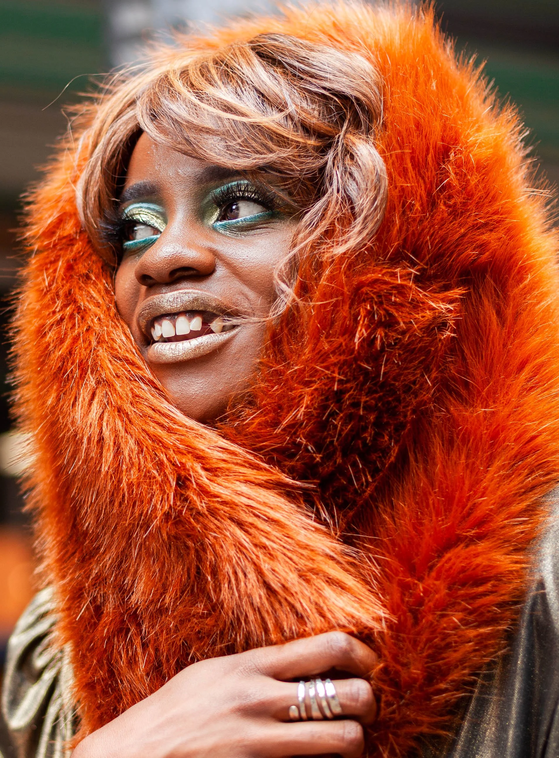 A woman with colorful makeup and shiny lips, wearing an orange furry coat, smiling and holding her hand with rings near her chest.