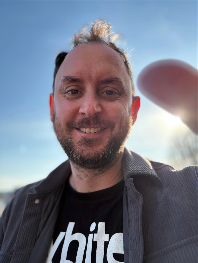A smiling man with a beard taking a selfie outdoors during daylight, with the sun behind him and a clear blue sky.