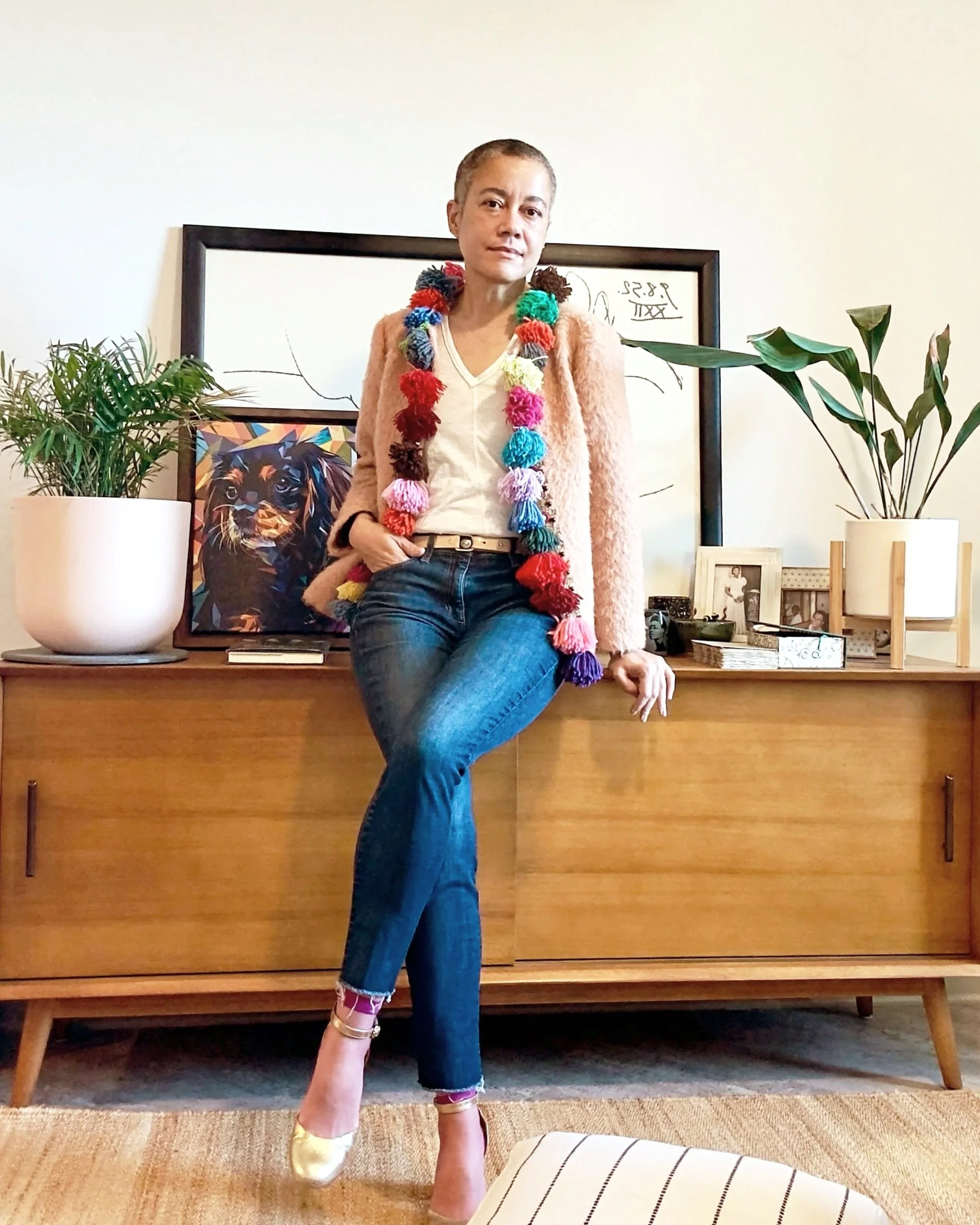 A woman standing in front of a wooden sideboard with plants, framed photographs, artwork, and decorative objects, wearing a colorful pom-pom-lined blazer, white blouse, jeans, and gold shoes.