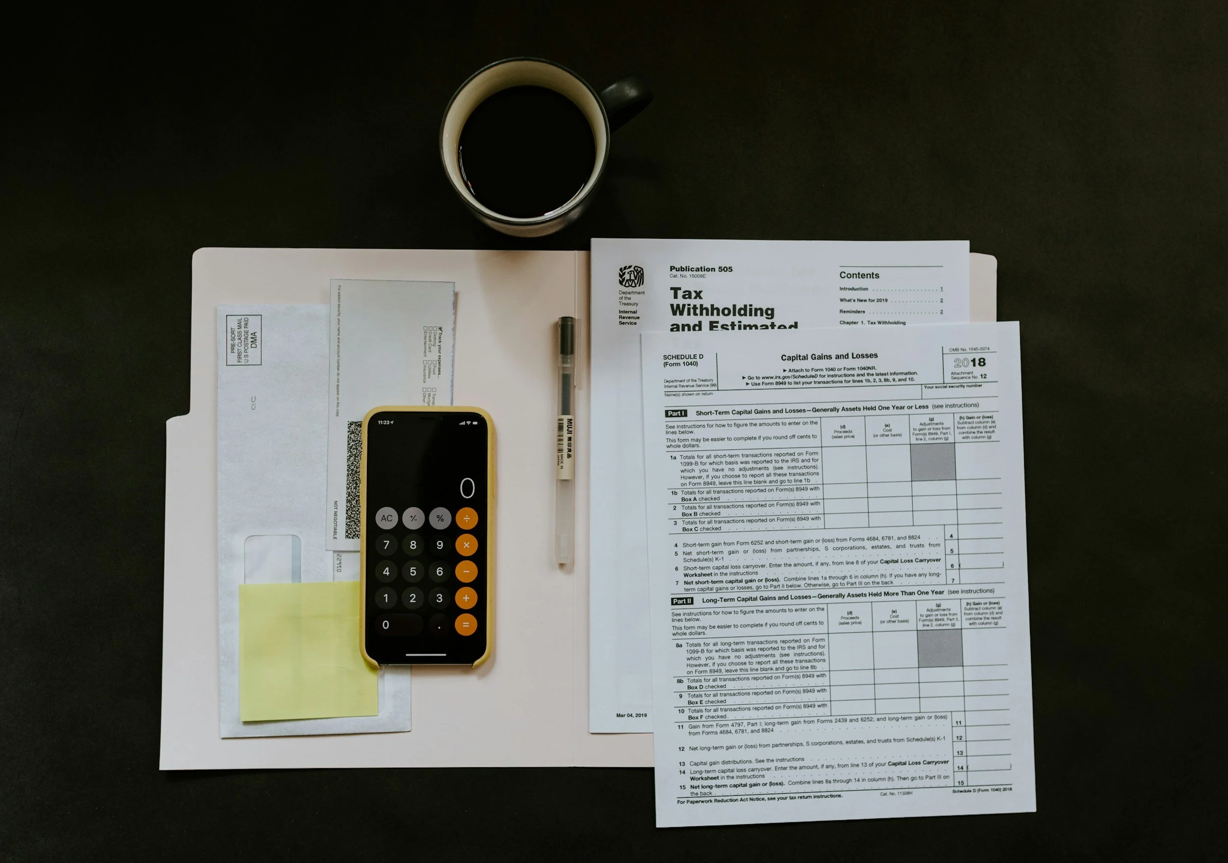 A desk with a cup of coffee, tax documents, a calculator on a sticky note pad, a pen, and a postal mail envelope.