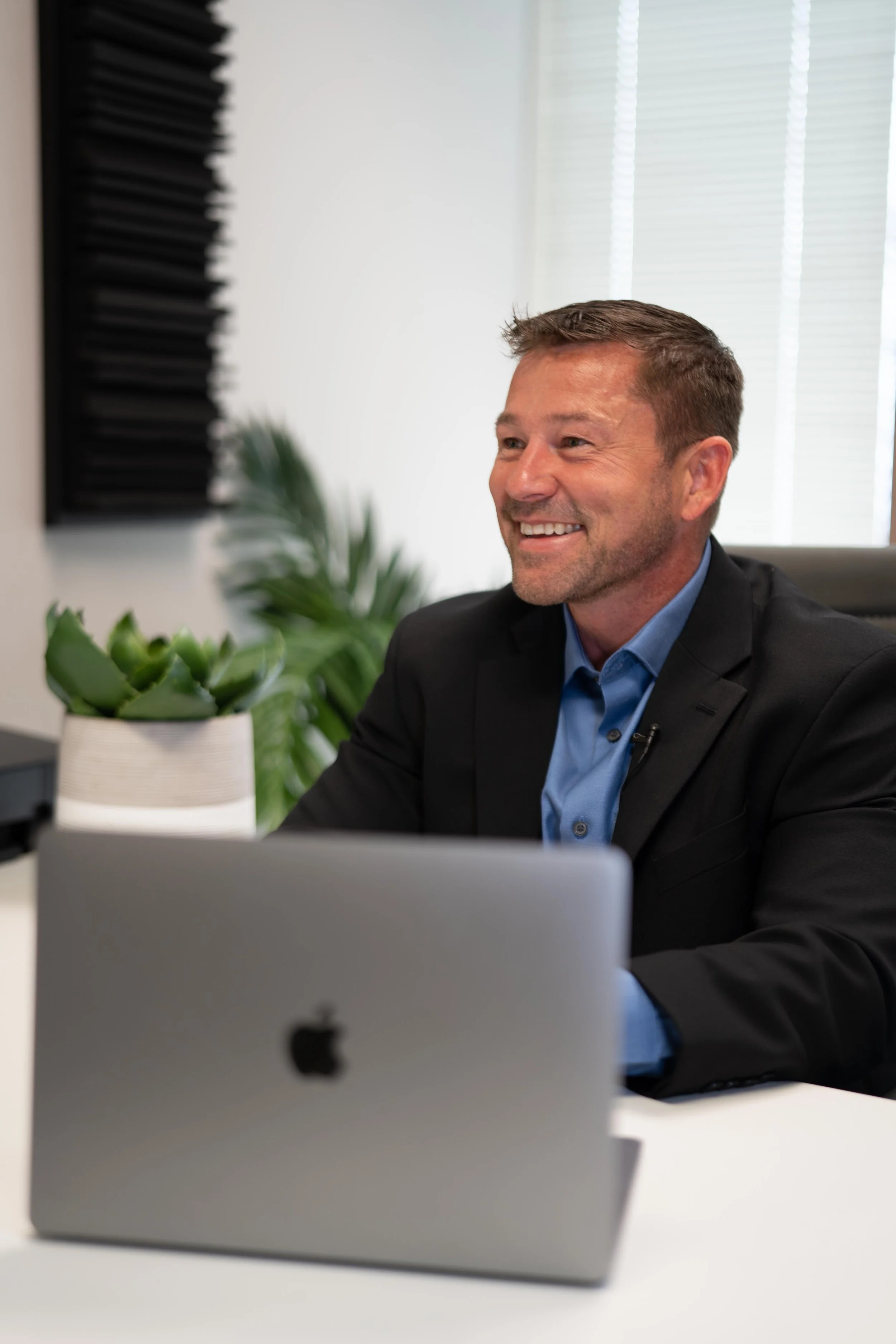 A smiling man in a suit sitting at a desk with a silver laptop and a potted succulent plant, in an office with blinds and a plant in the background.