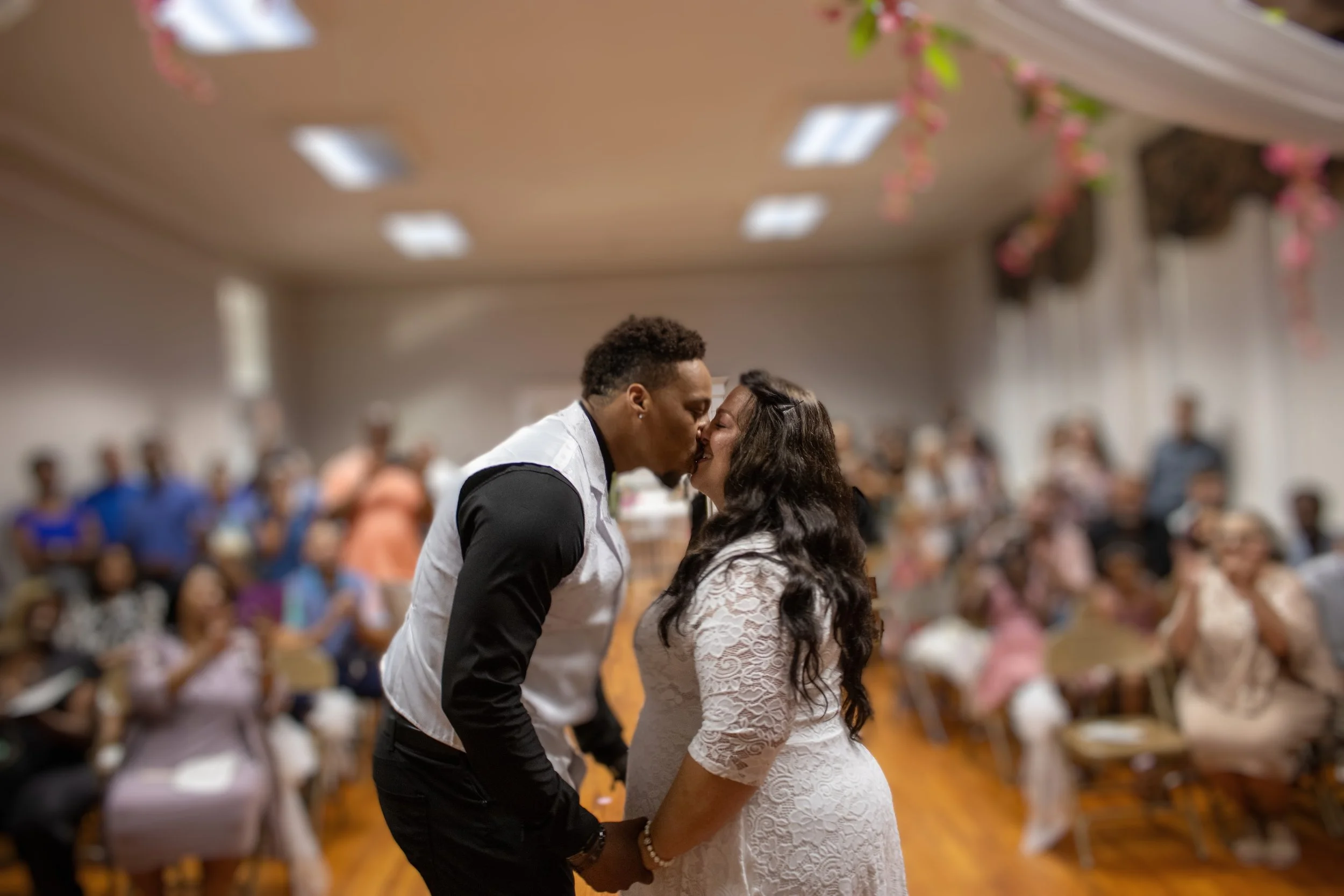 A couple kissing during their wedding reception with guests seated at tables in the background.