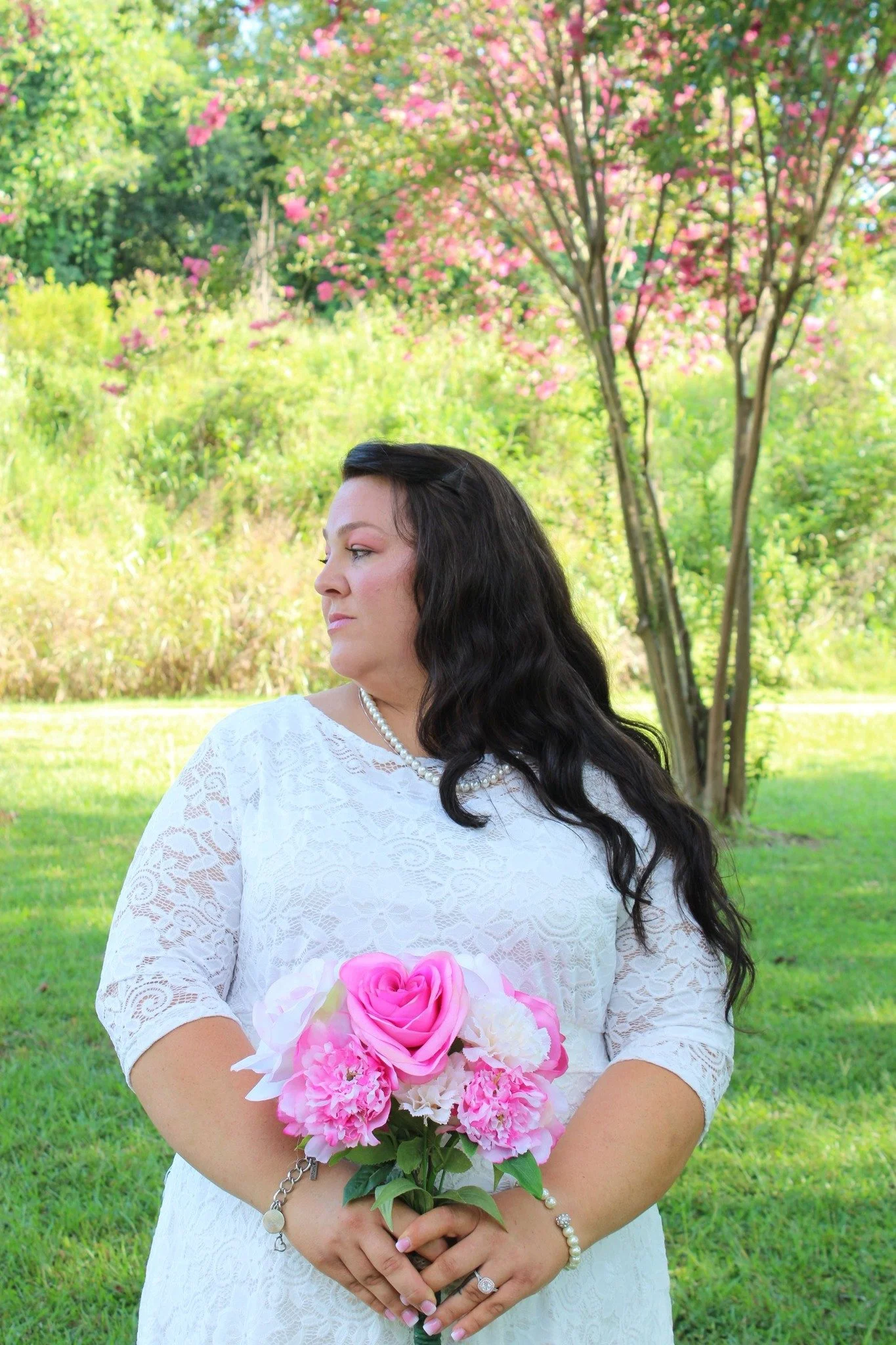 A woman with long dark hair, wearing a white lace dress and a pearl necklace, is holding a bouquet of pink and white flowers, standing outdoors near a flowering tree with pink blossoms.