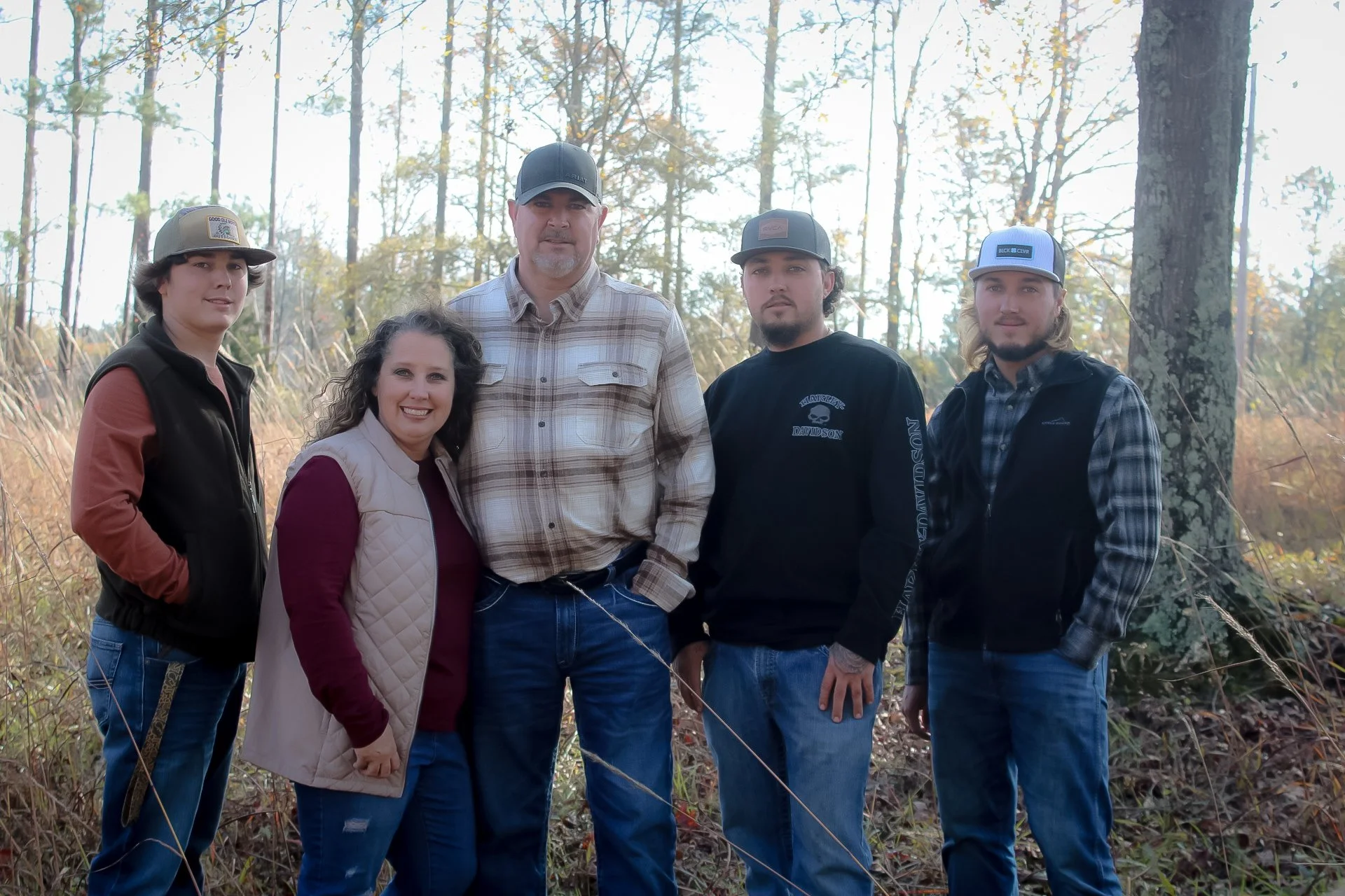 A group of five people standing outdoors in a wooded area during daytime, smiling for a photo, dressed in casual and outdoor clothing.