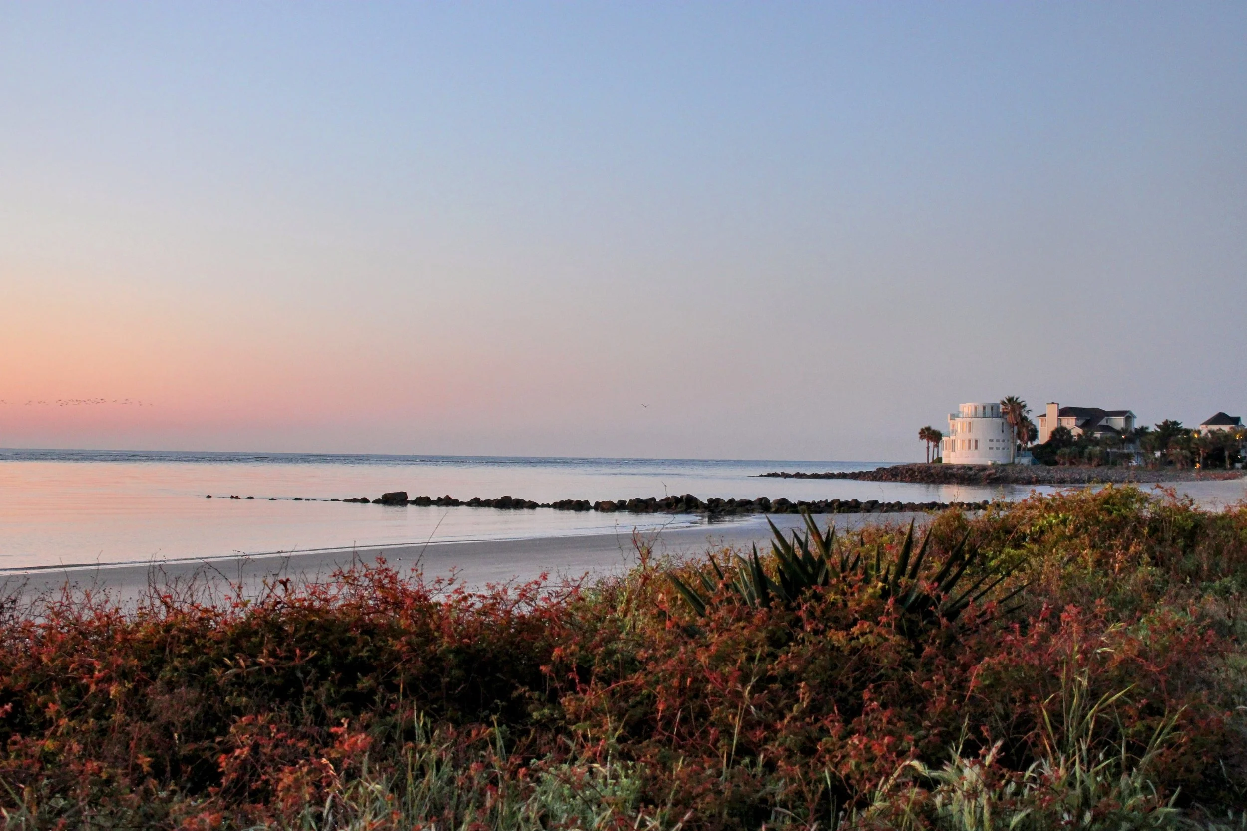 Sunset over a beach with a house on a small pier, rocks in the water, and bushes in the foreground.