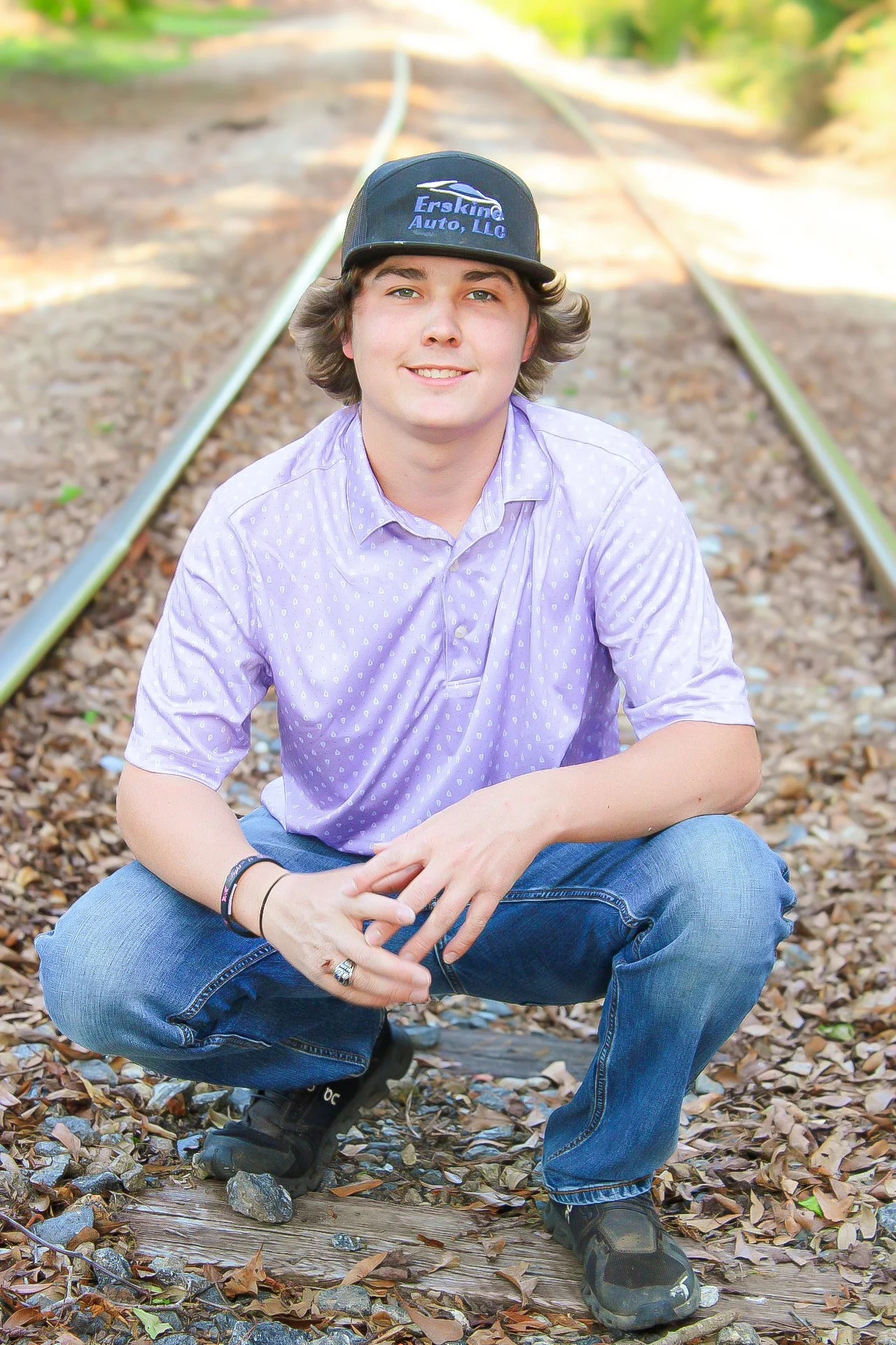 A young man in a purple patterned shirt kneeling on an old railroad track surrounded by fallen leaves, smiling at the camera with a natural background of trees and sunlight.