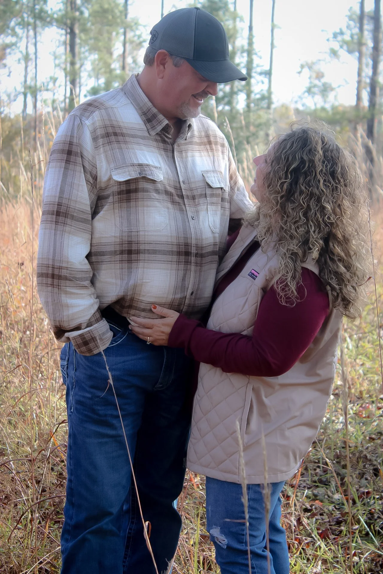 A man and woman stand close outdoors in a forested area, looking into each other's eyes and smiling, with the woman's arms around the man's waist.