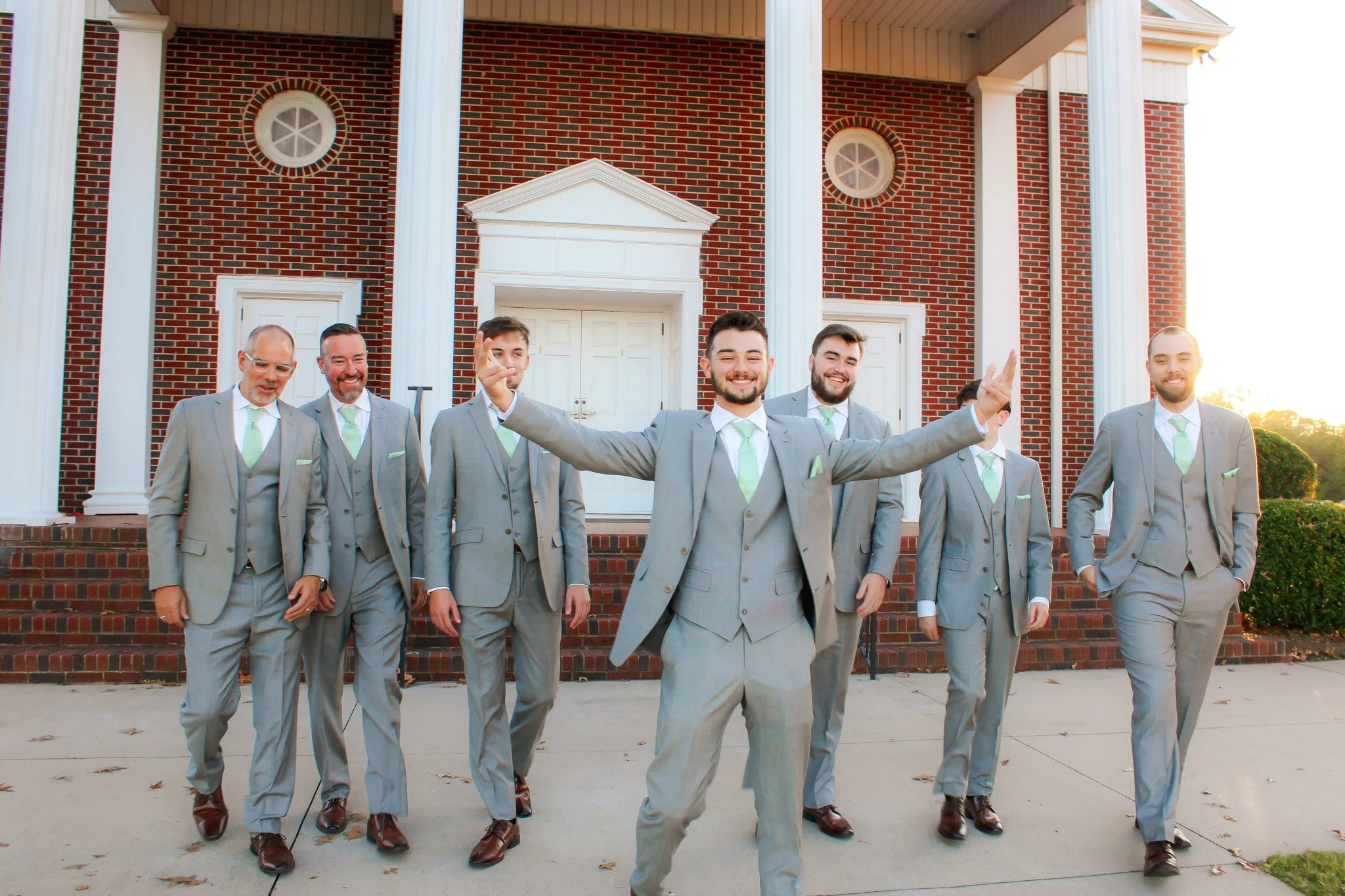 A group of seven men dressed in light gray suits with mint green ties walking out of a brick building with white columns and steps, smiling and posing for a photo.