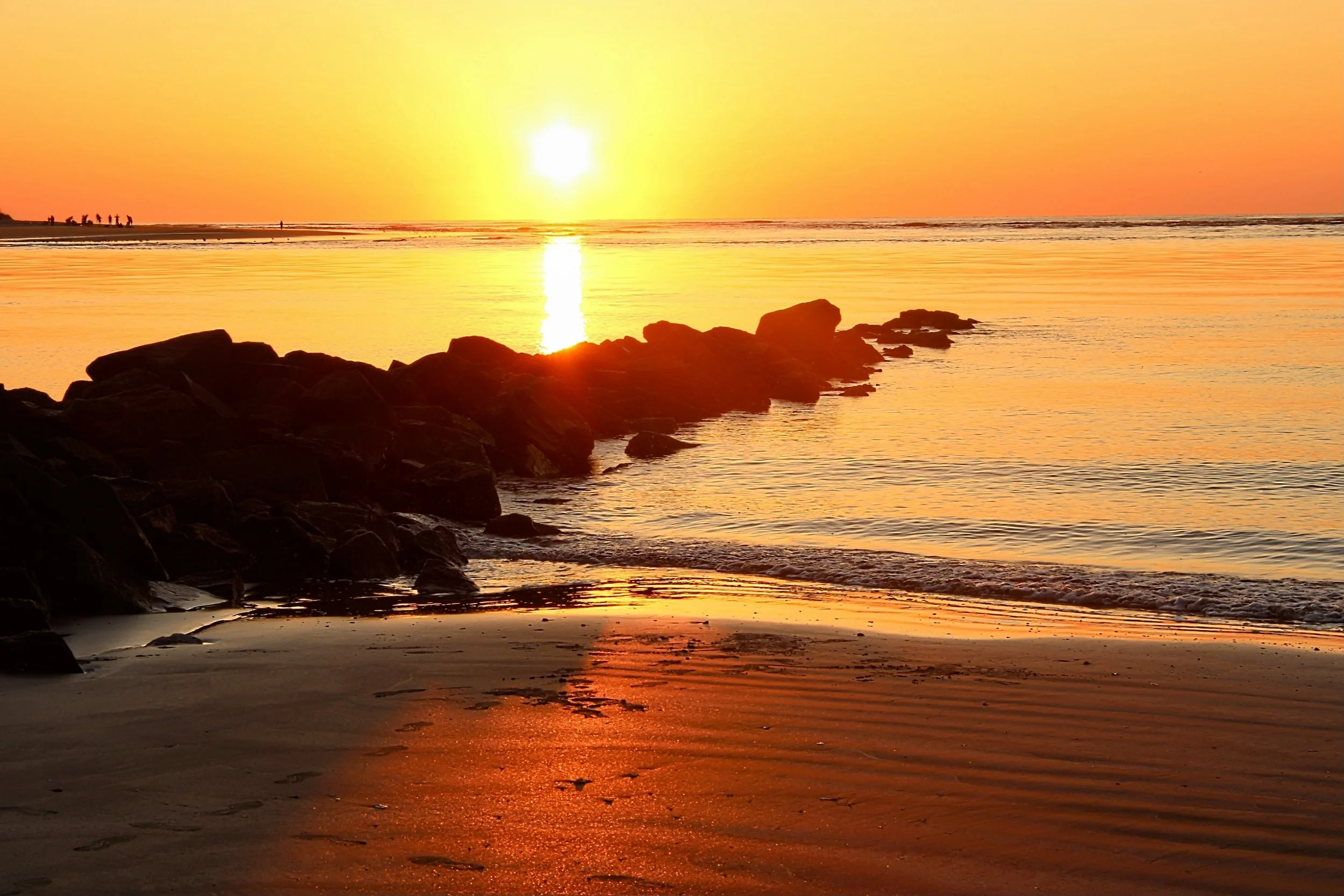 Sunset over a beach with rocks in the foreground and a group of people in the distance