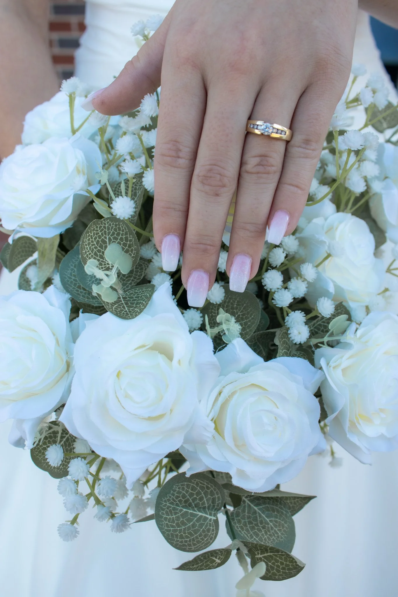 A person's hand with a diamond engagement ring on finger, resting on a bouquet of white roses and small white filler flowers, with green leaves, held against a blurred outdoor background.