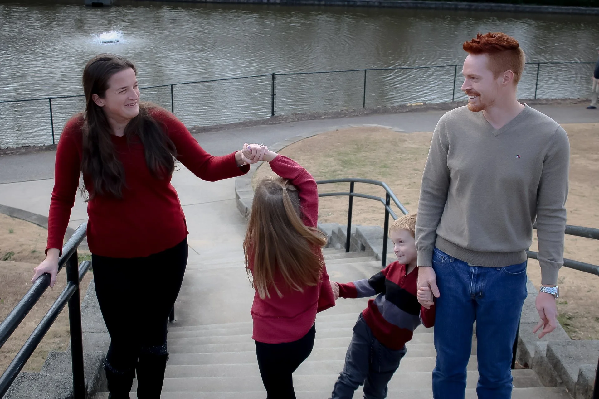 A family of four, two adults and two children, are holding hands and smiling on a set of outdoor stairs near a body of water.