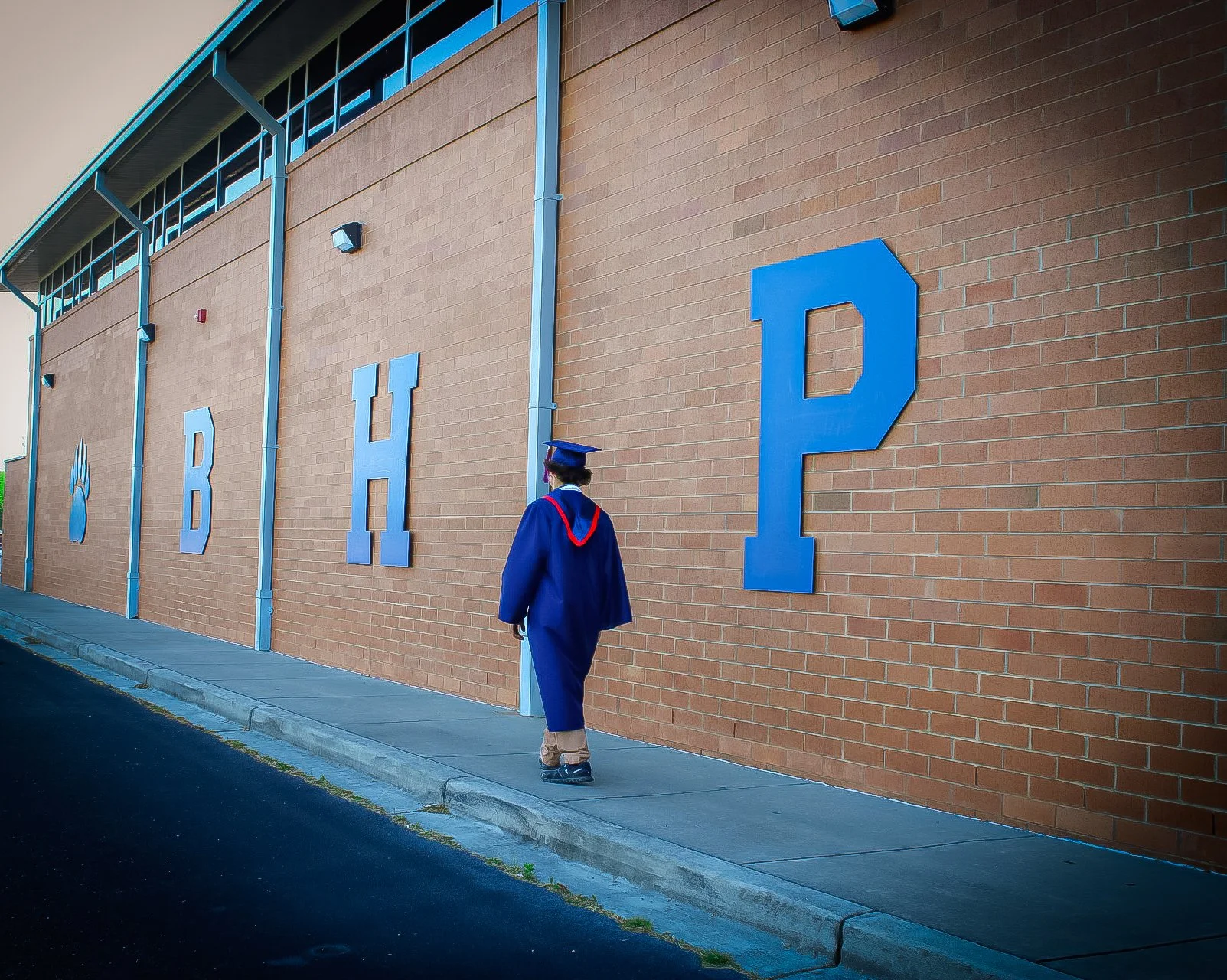 Person in a graduation gown and cap walking past a brick school building with large blue letters spelling "BHD P" on the wall.