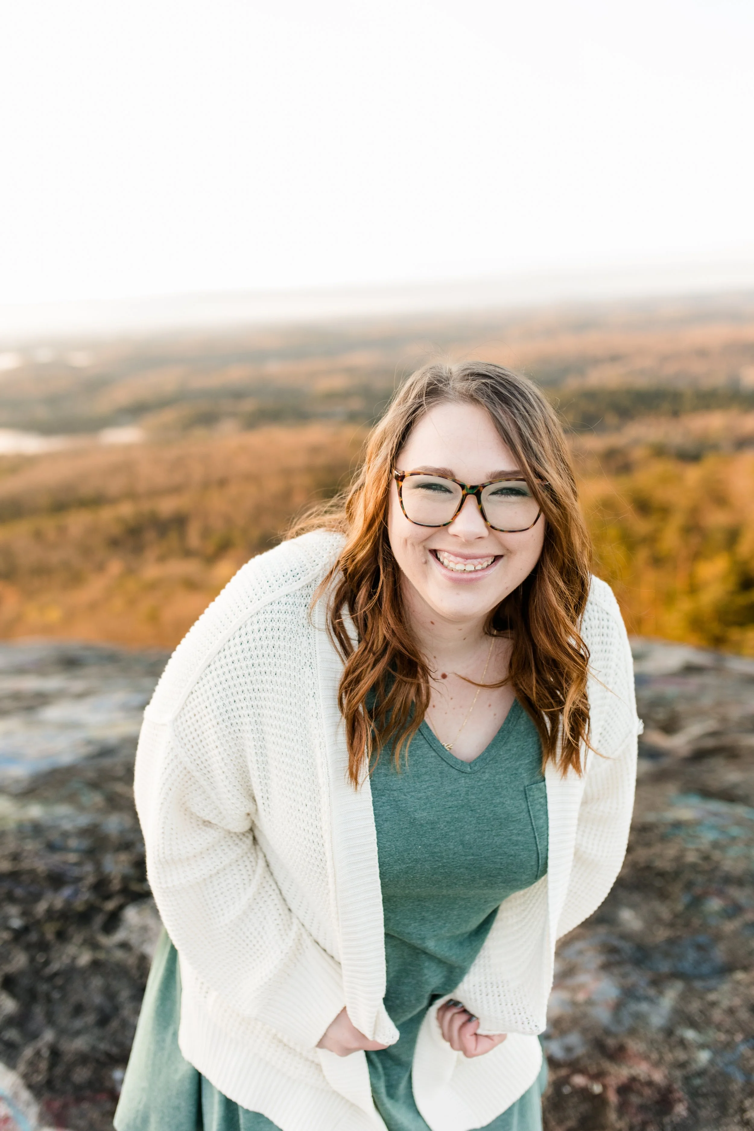 Smiling young woman with glasses in a green shirt and white sweater, outdoors with autumn-colored trees and a distant horizon in the background.