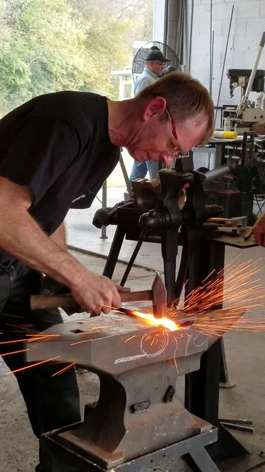A man is blacksmithing, striking metal on an anvil with sparks flying as he hammers. Another person is working in the background near tools and equipment.