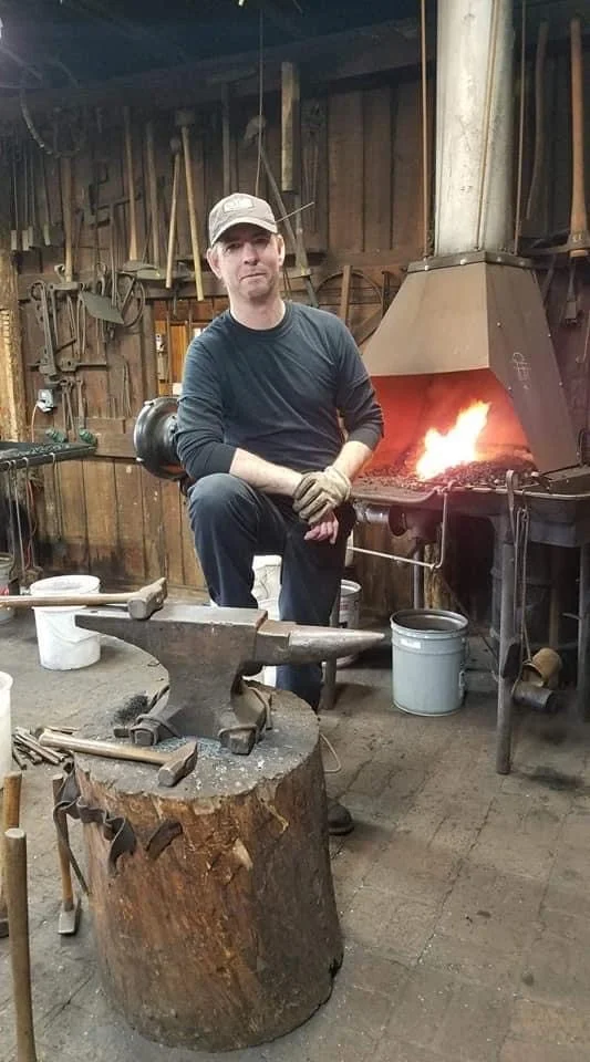 Blacksmith in a workshop holding a heated piece of metal. The workshop has tools hanging on wooden walls, an anvil on a tree stump, and a forge with a fire glowing.