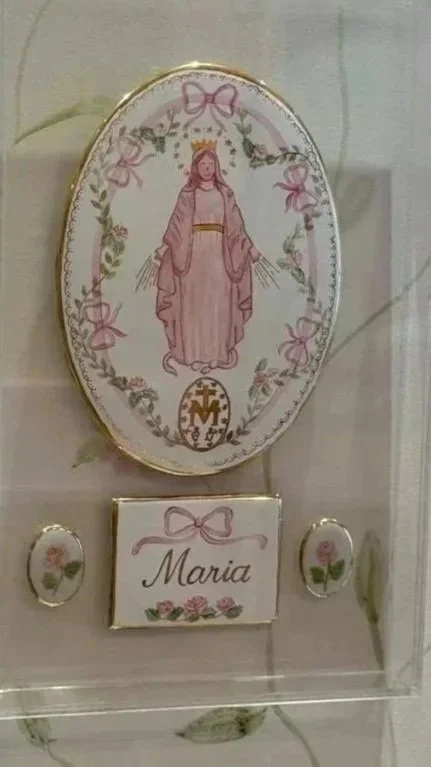 Imagem de uma placa decorativa com uma ilustração de Maria, uma cruz e a palavra 'Maria' escrita, decorada com rosas e fitas.