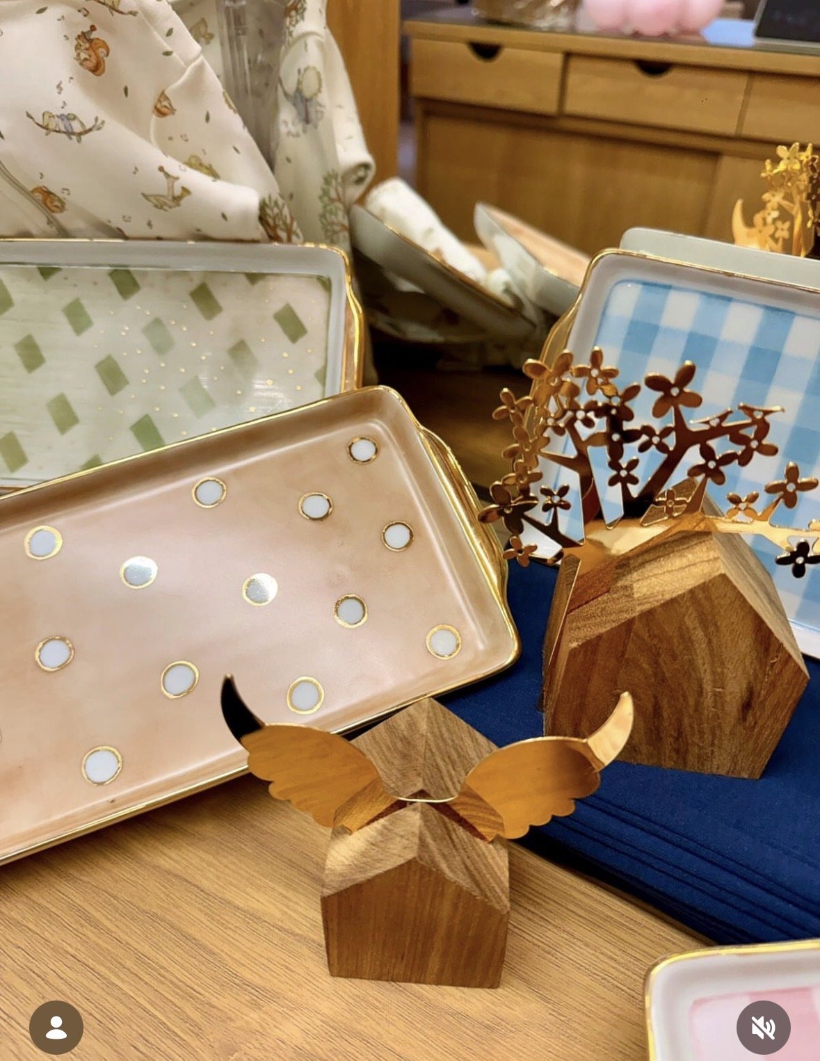 Vários objetos decorativos em tons de dourado e madeira, incluindo bandejas, suportes e uma árvore decorativa com moldura de madeira, dispostos em uma mesa de madeira.