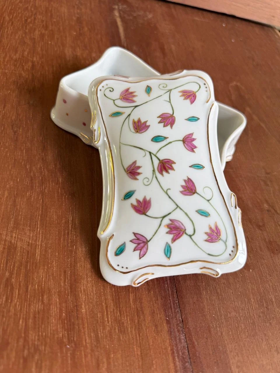 Porta objetos de porcelana branco com detalhes em ouro, decorado com flores rosa e folhas azul e verde, em formato de caixa retangular com curvas.