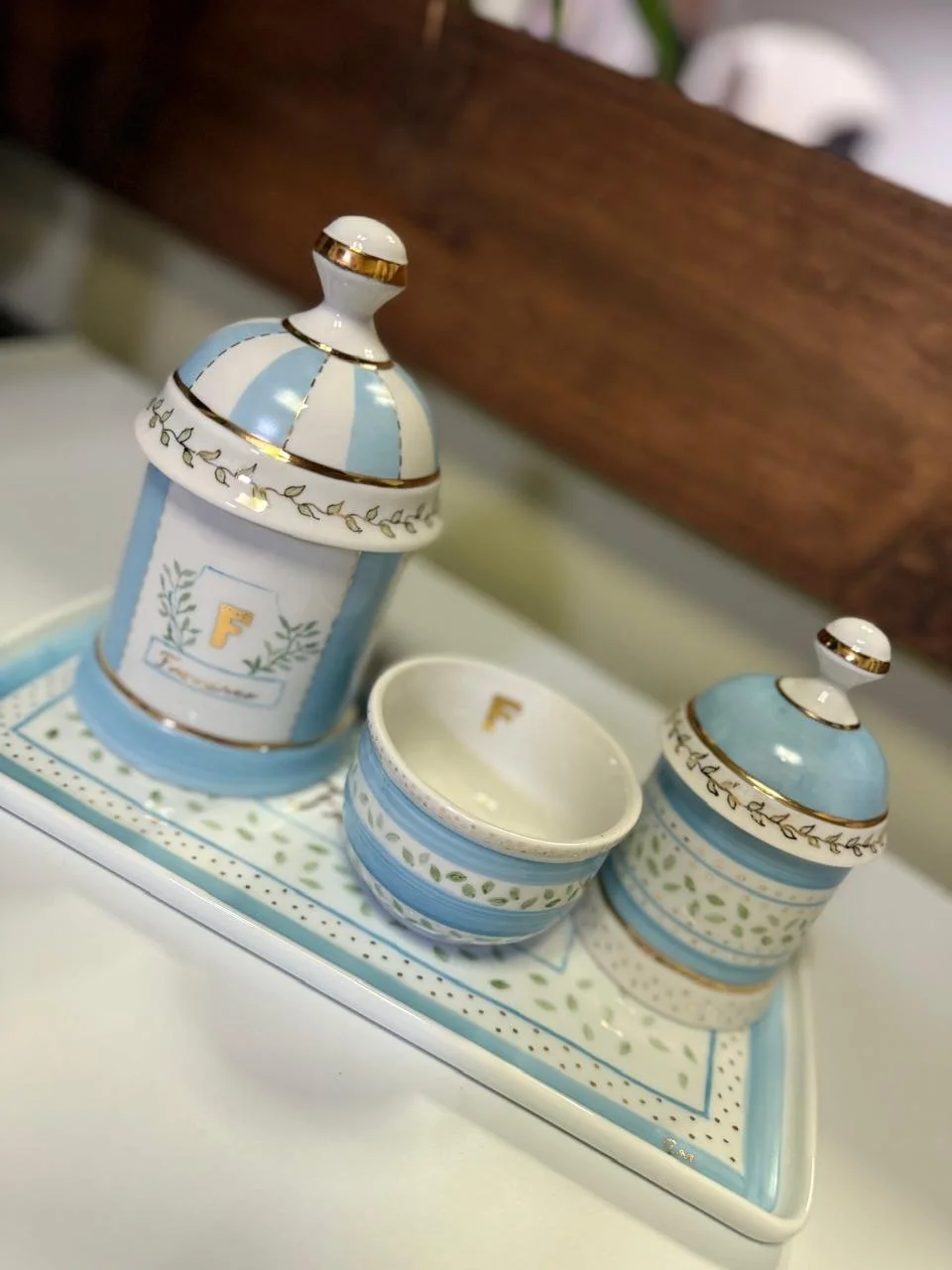 Conjunto de potes decorativos com tampas, azul e branco, com detalhes dourados, colocados em uma bandeja decorativa com padrão de folhas e pontos