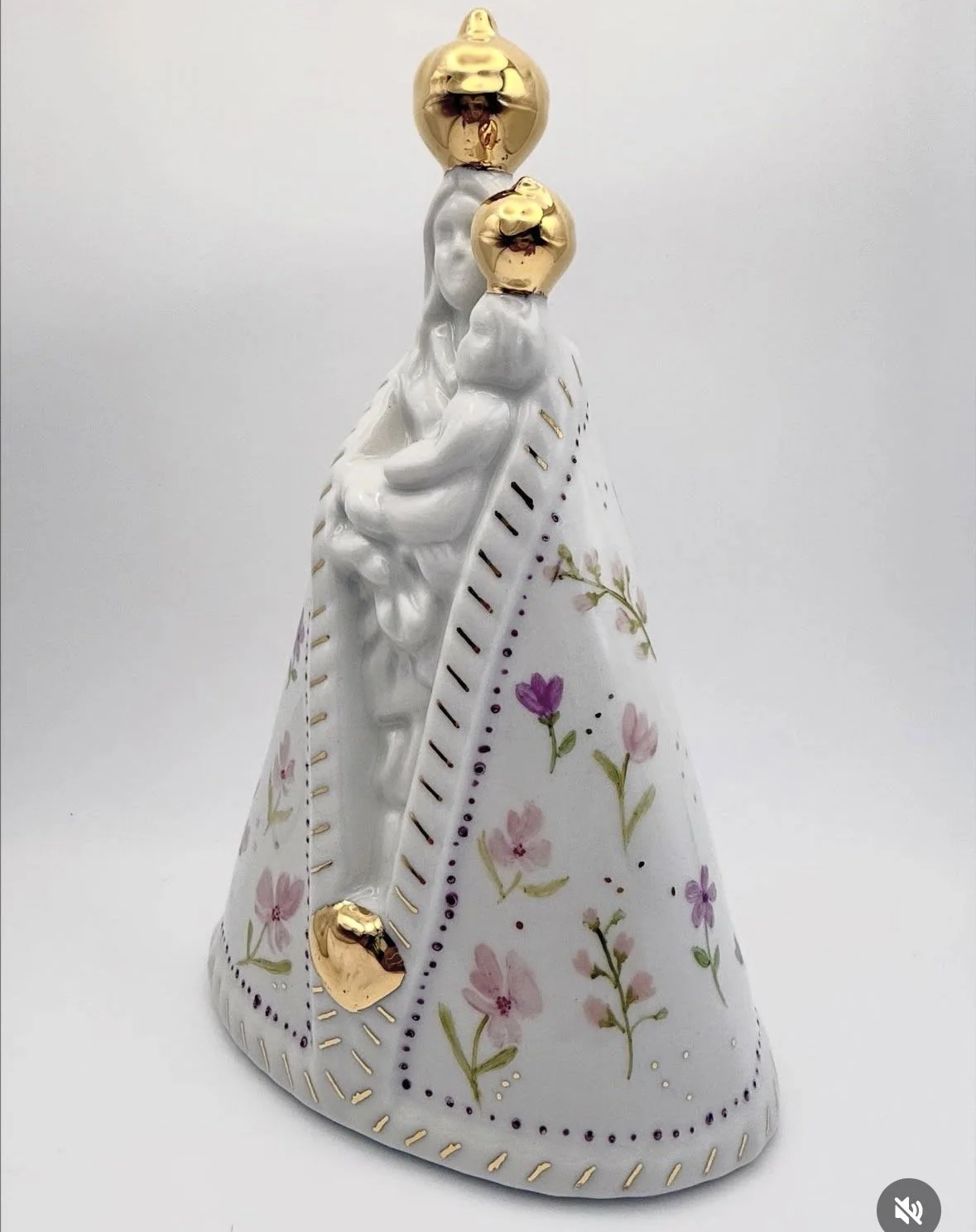 Imagem de uma escultura de porcelana decorativa que representa uma figura feminina com uma capa floral, com detalhes dourados, e cabeças douradas em estilo abstracto.