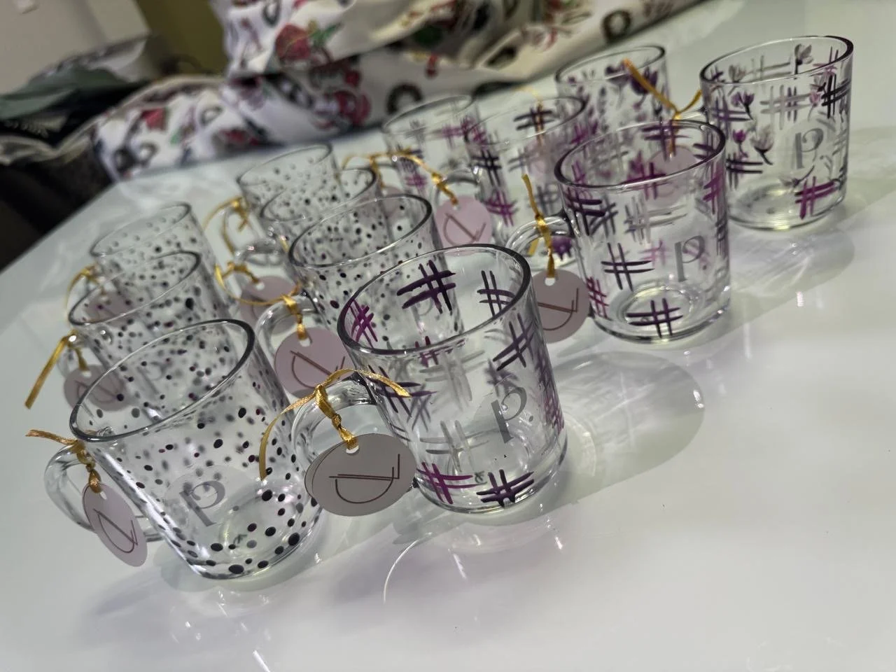 Conjunto de 12 canecas de vidro decoradas com marcas de hashtag e bolinhas, algumas com tags de presente penduradas.