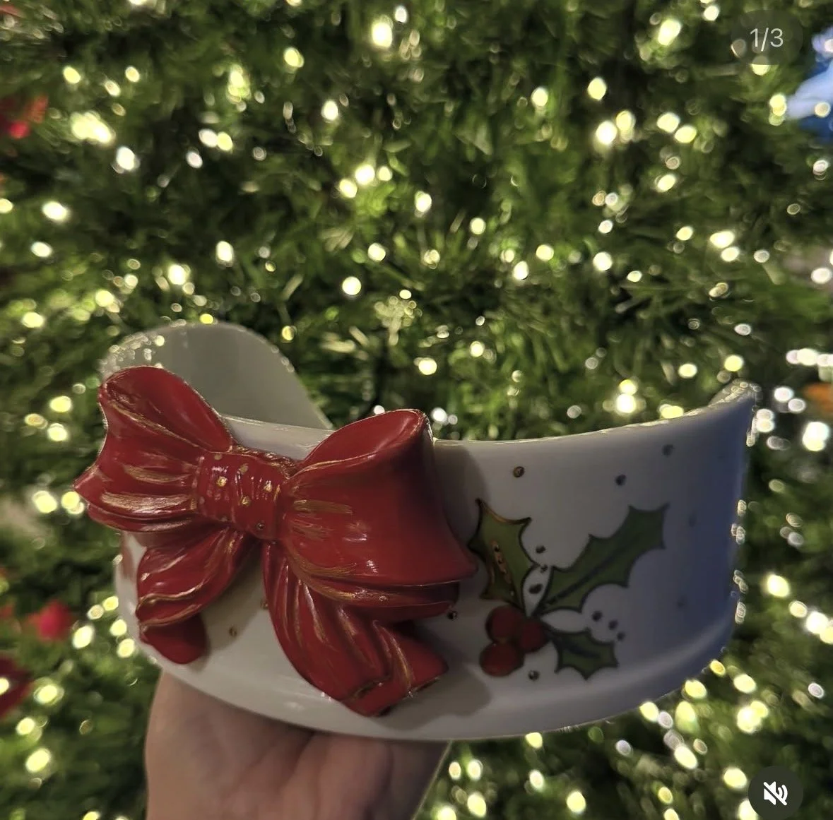 Xícara decorada com laço vermelho e folhas de azevinho, em primeiro plano, com árvore de Natal iluminada ao fundo.