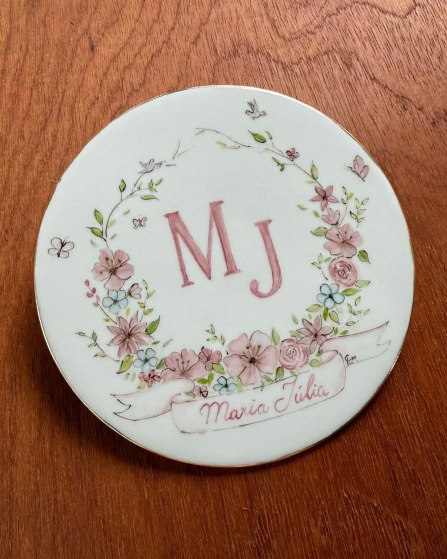 Prato decorado com o monograma 'M J', cercado por uma coroa de flores cor-de-rosa e azuis, com pássaros, borboletas e uma faixa escrita 'Maria Júlia'.