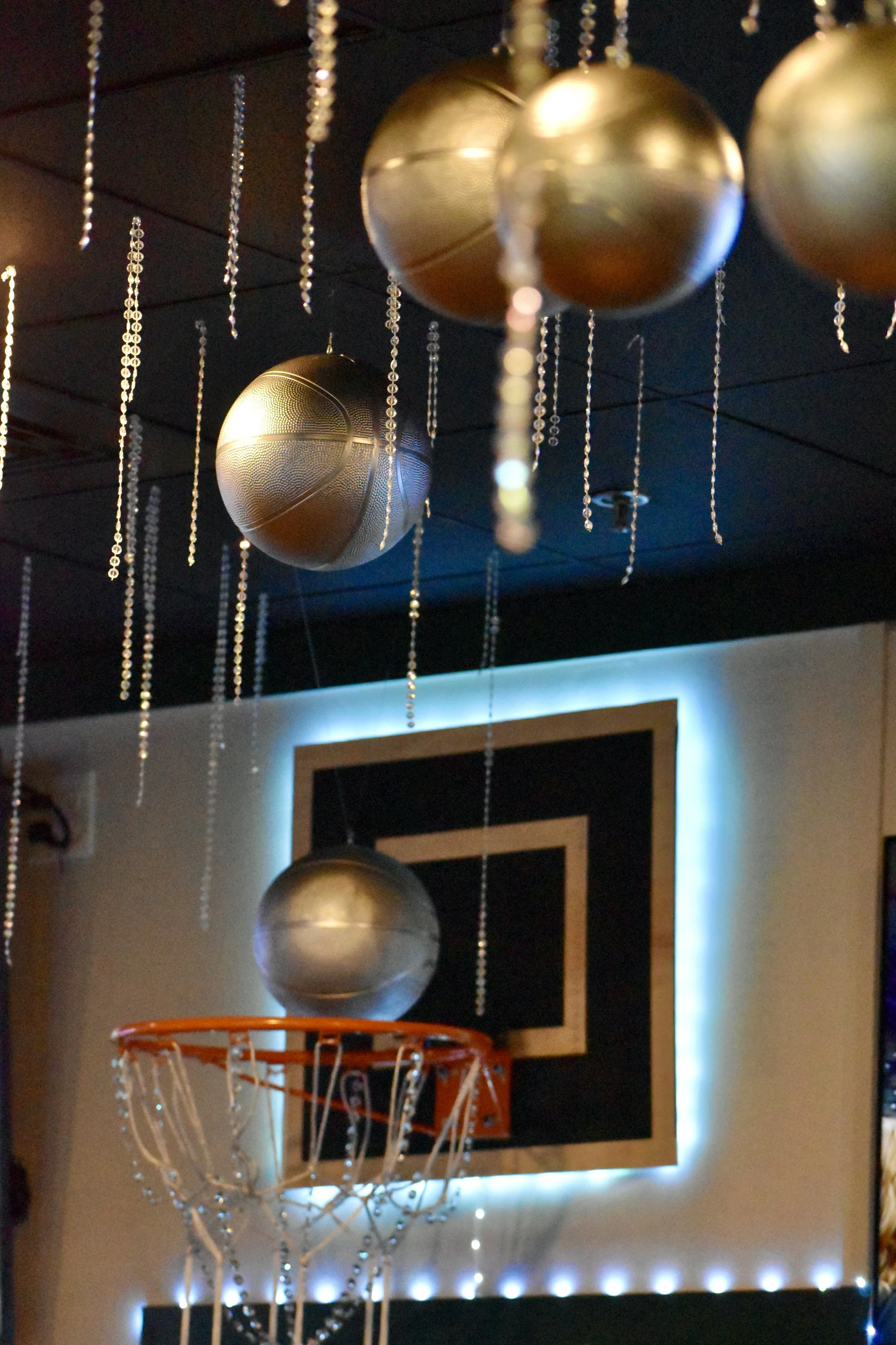 Indoor basketball court with a backboard and a hoop, decorated with hanging metallic balls and string lights. March Madness