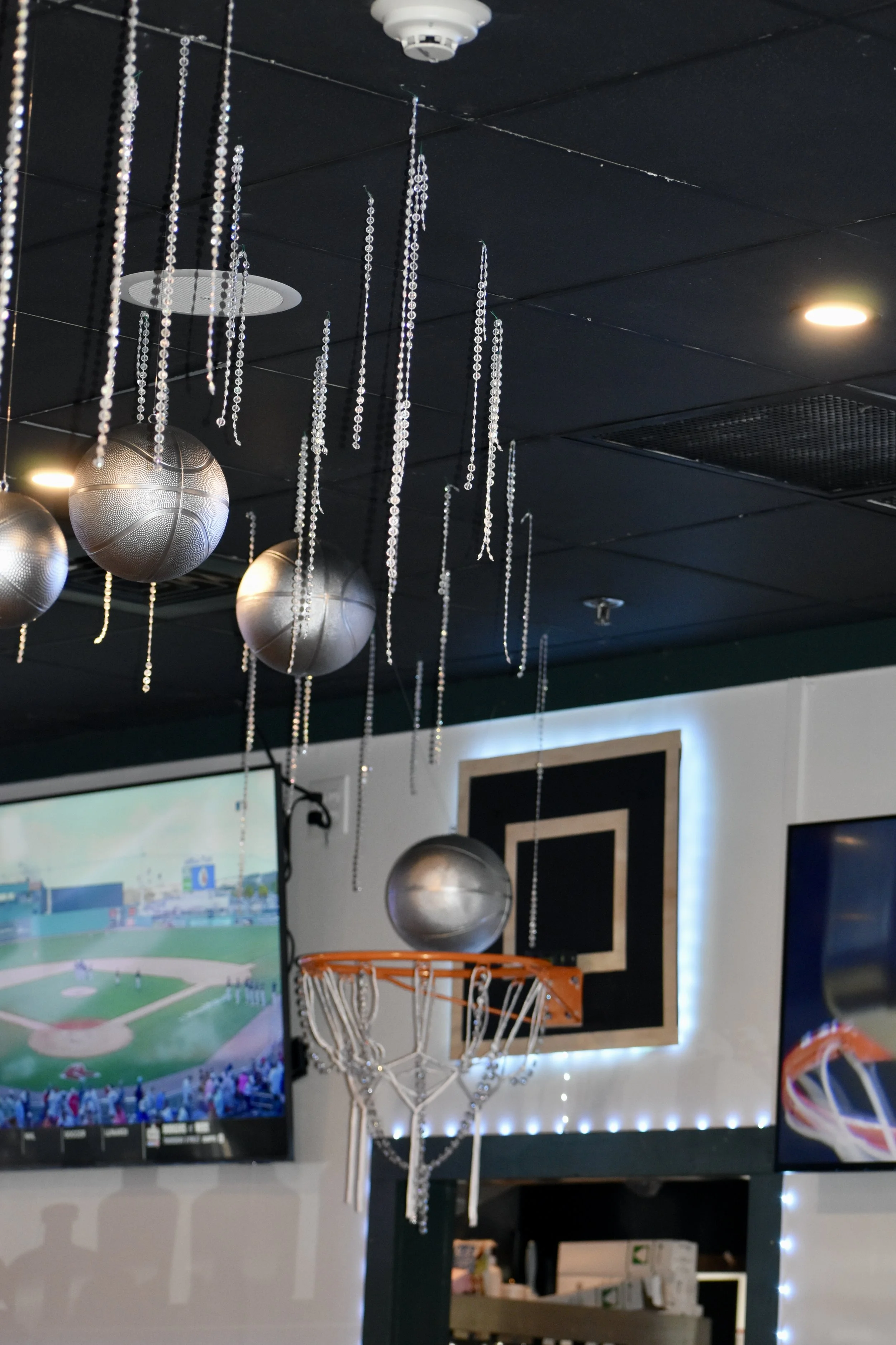 Indoor sports bar with decorative hanging basketballs, silver gym balls, a basketball hoop with beads, and sports screens. March Madness