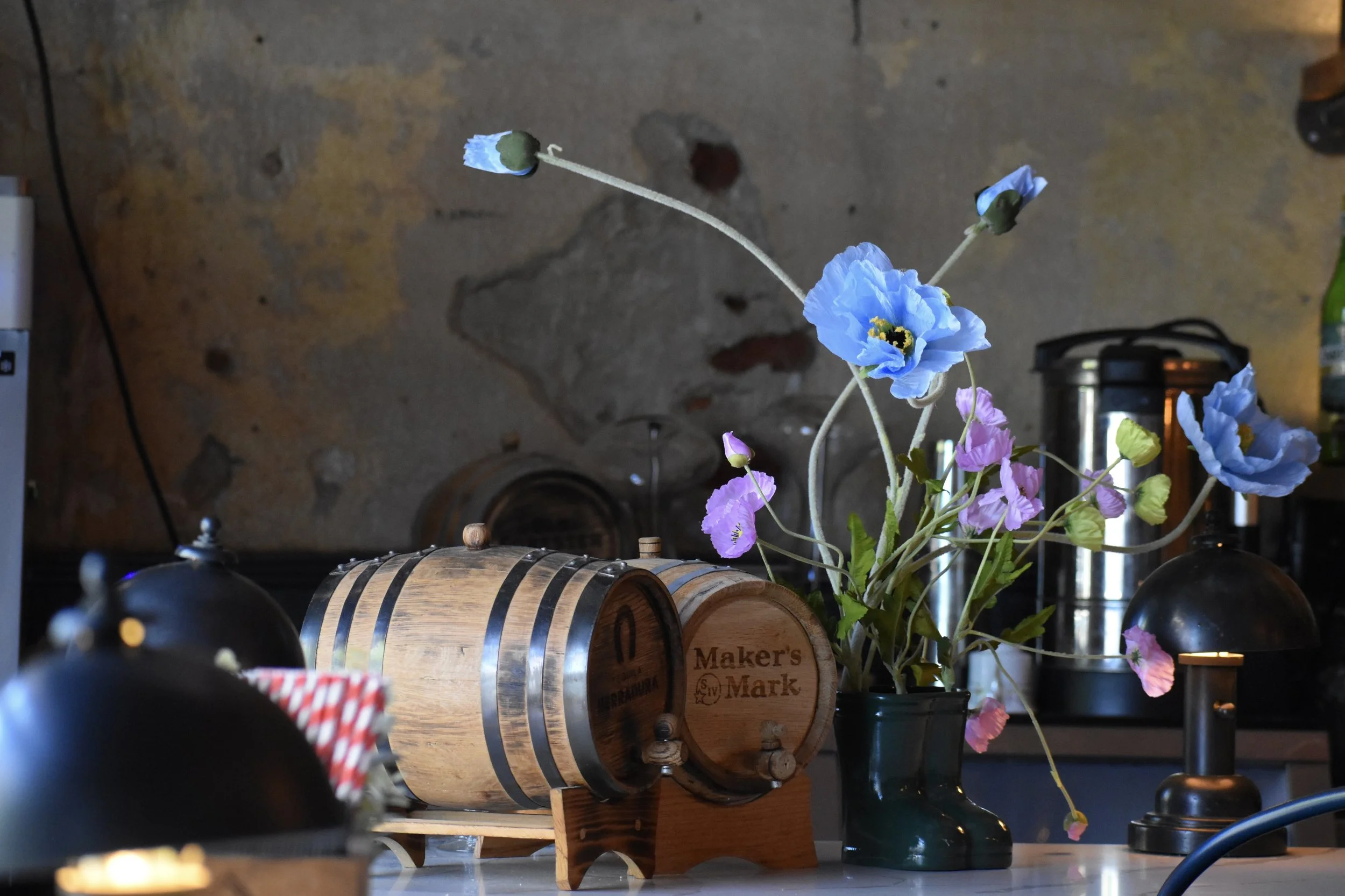 Decorative scene with a small wooden barrel, a flower vase with blue and pink flowers, and some black lamps on a table against a textured concrete wall.
