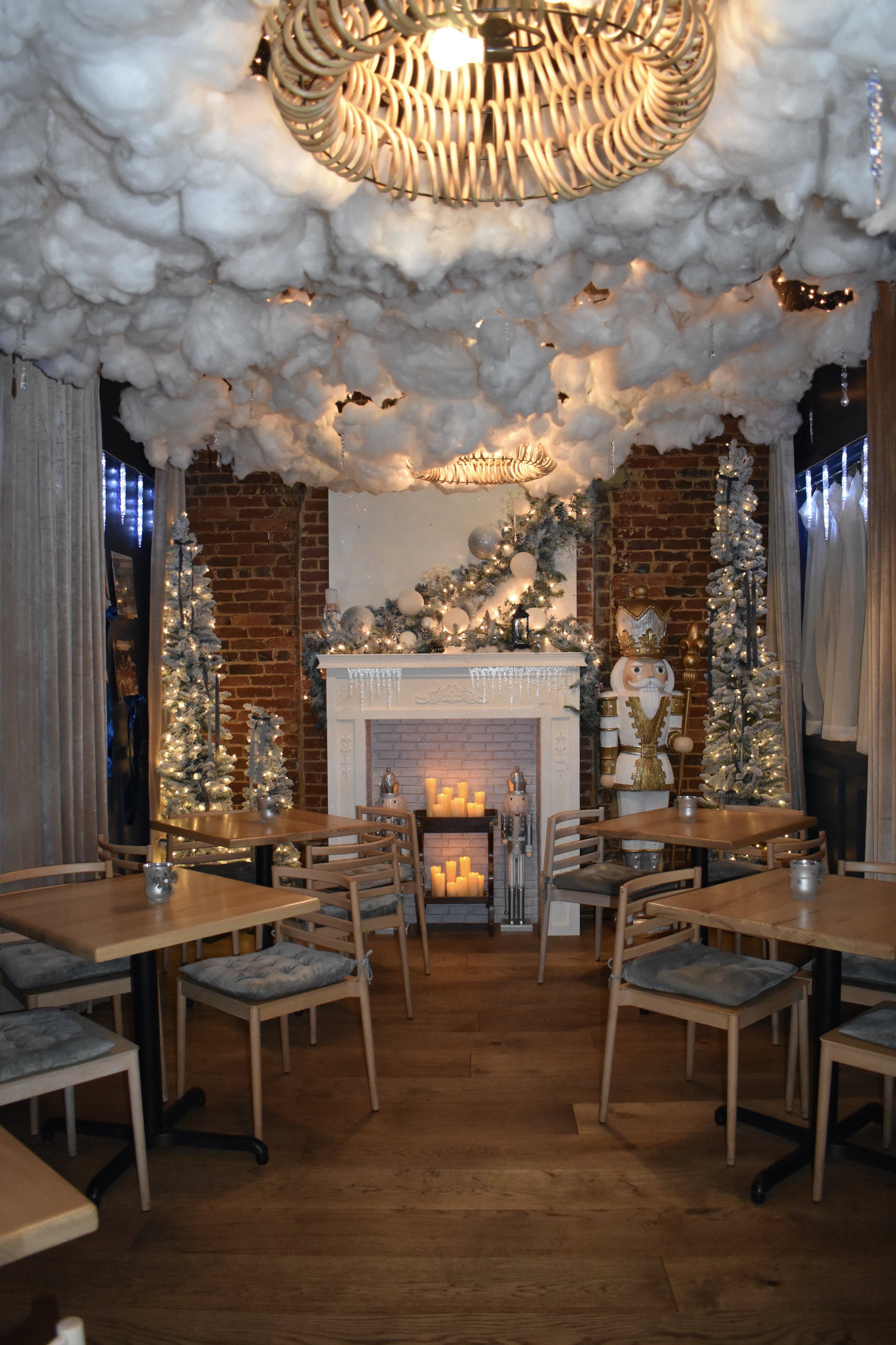 A cozy room decorated for Christmas with a fireplace, lit candles, white Christmas trees, festive garlands, and Nutcracker figures. The ceiling is decorated with cotton clouds and hanging lights, creating a magical holiday atmosphere.