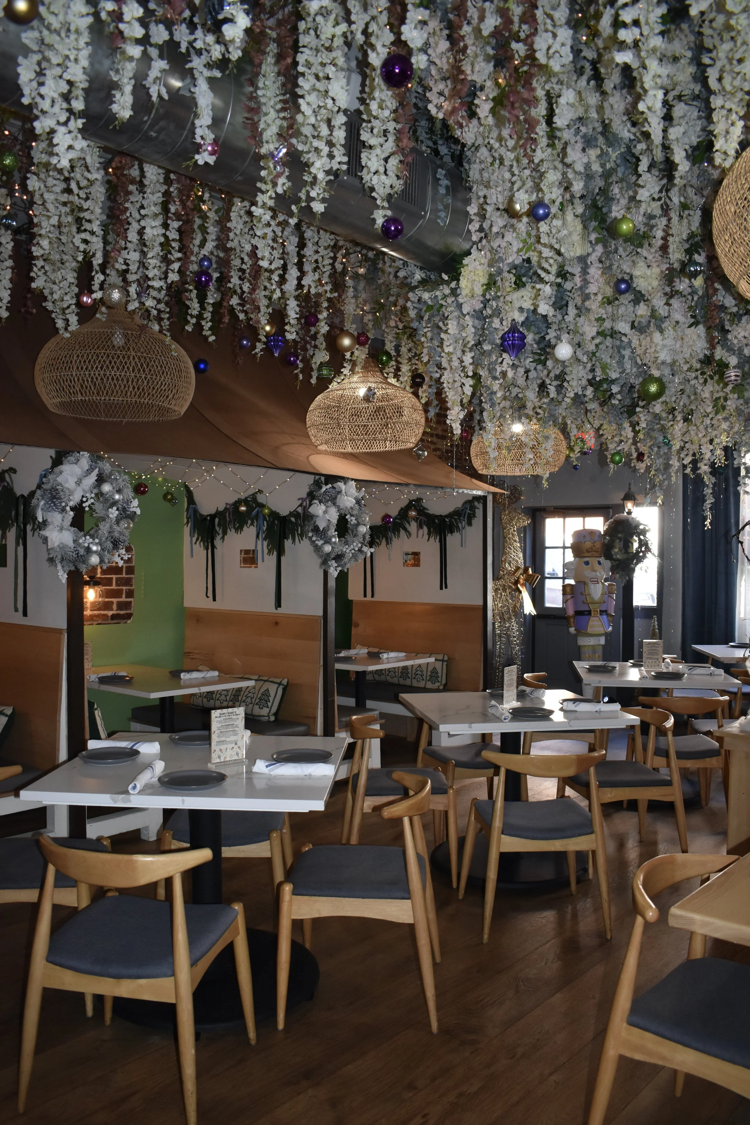 Interior of a restaurant decorated for the holiday season with white flowers hanging from the ceiling, colorful ornaments, wicker pendant lights, and holiday wreaths and garlands on the walls.