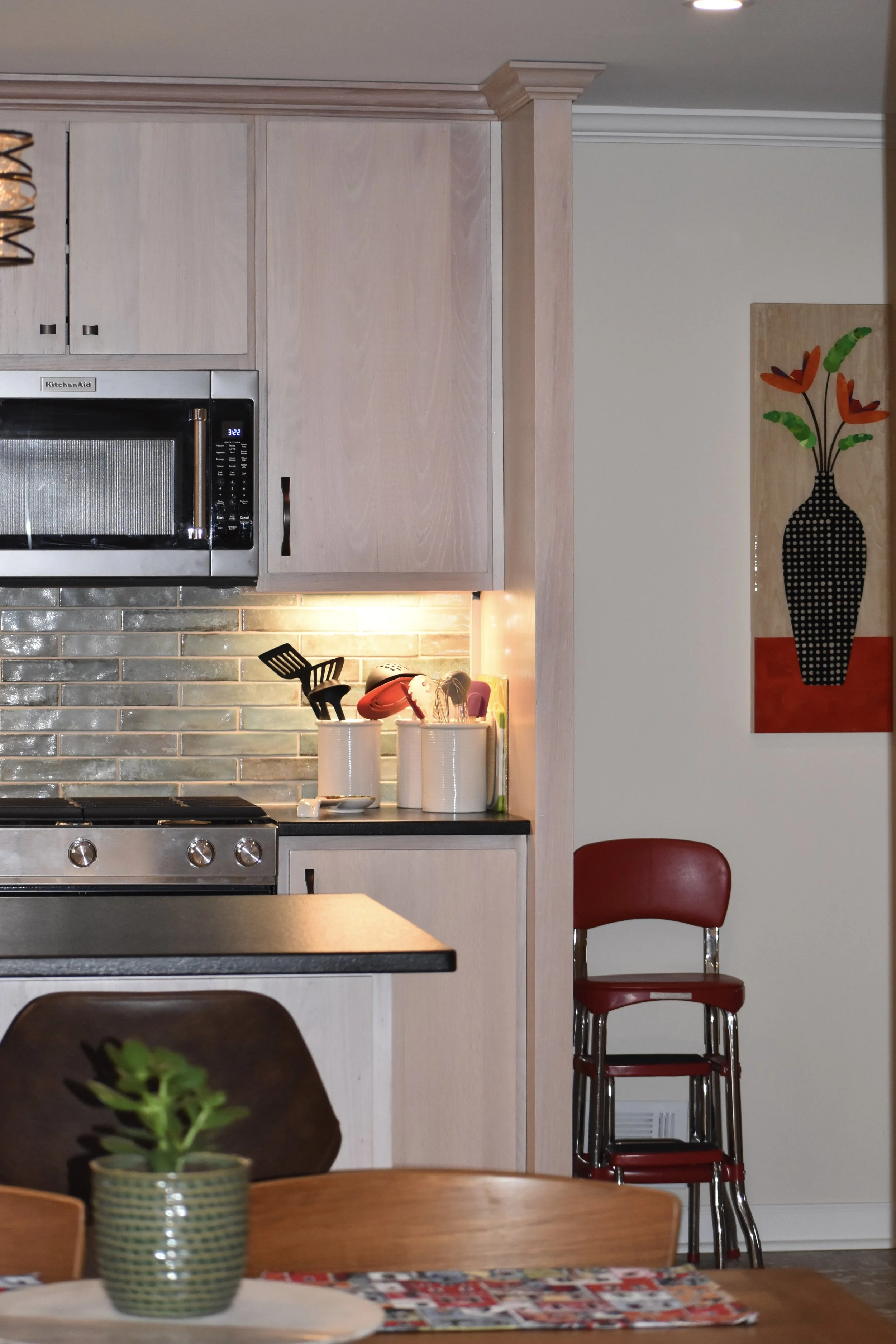 Part of a kitchen with light-colored cabinets, a microwave oven, a backsplash with gray bricks, and countertop items in white containers. In the foreground, a part of a dining table with a plant in a green pot and a wooden chair. To the right, a red 