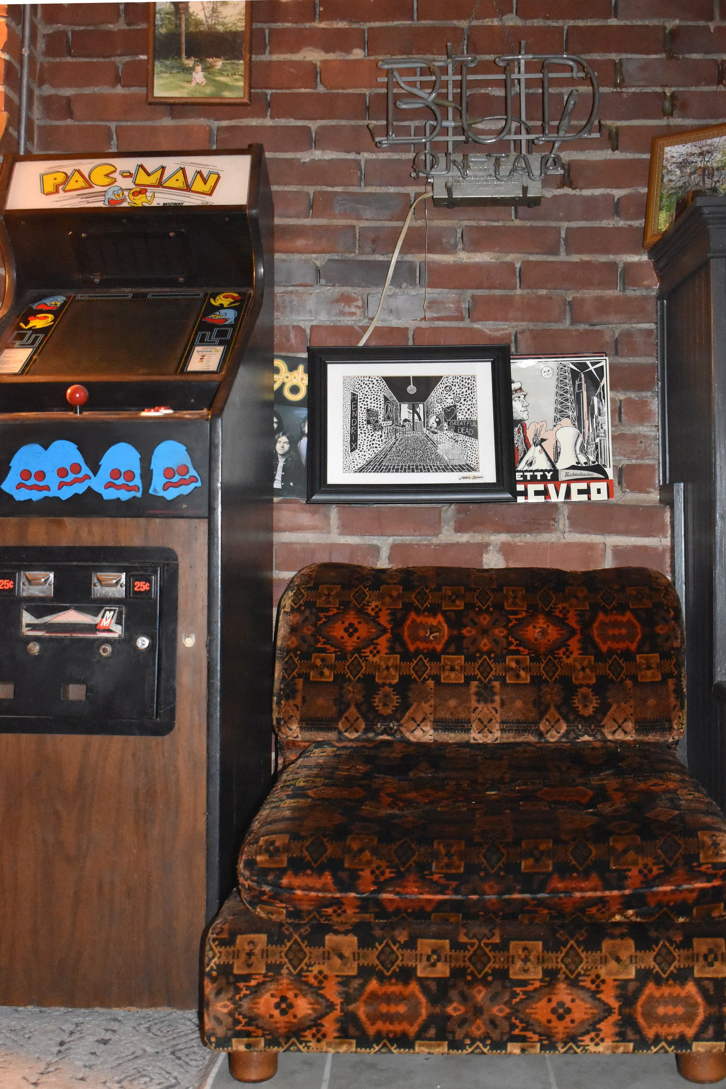 A vintage arcade game machine next to a patterned upholstered chair against a brick wall with framed artwork and a metal "BED" sign.