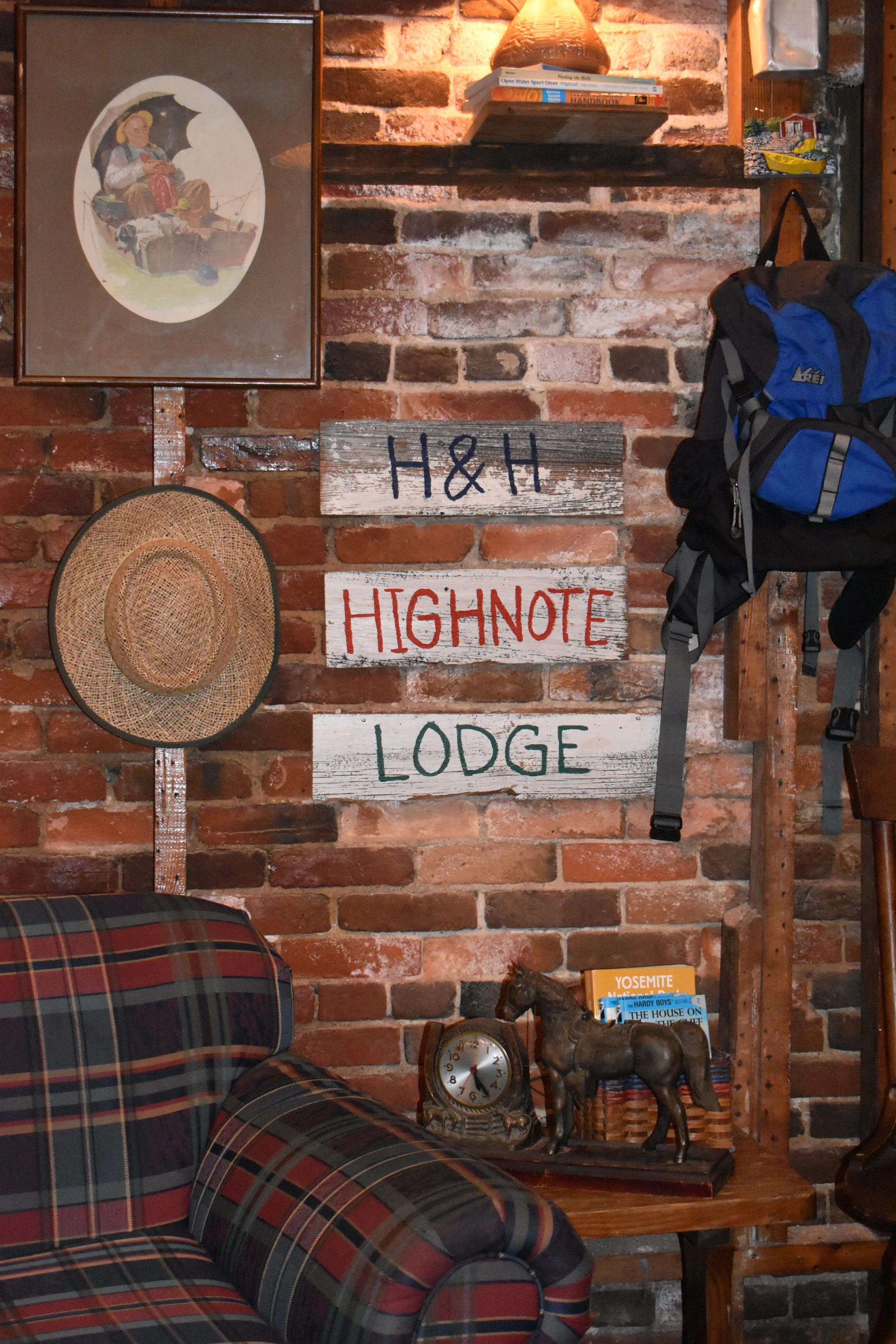 A cozy room with a brick wall decorated with a framed picture, a woven hat, and a wooden sign reading "H&H HIGHNOTE LODGE." There is a plaid armchair in the foreground, a small wooden side table with books and a horse figurine, and a blue backpack ha