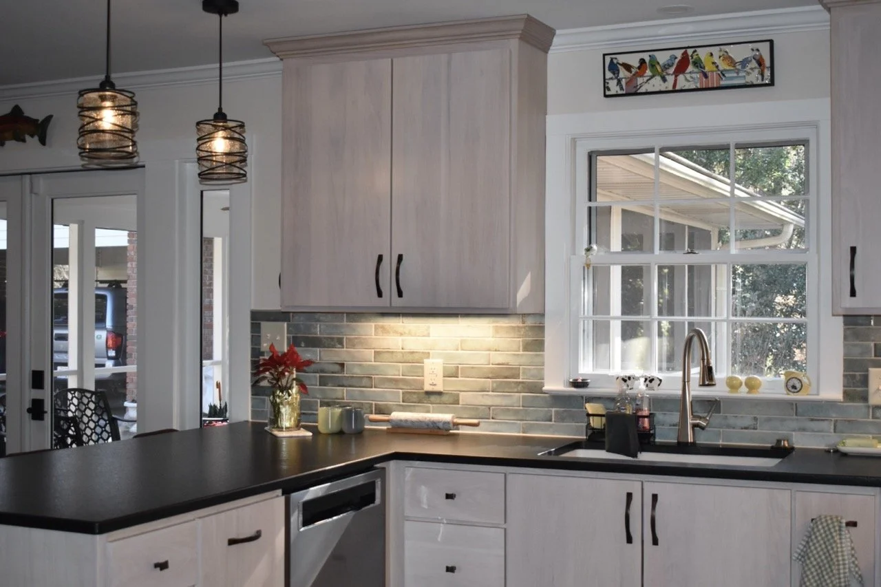 Kitchen with white cabinets, black countertop, green backsplash tiles, window above the sink, and hanging pendant lights.