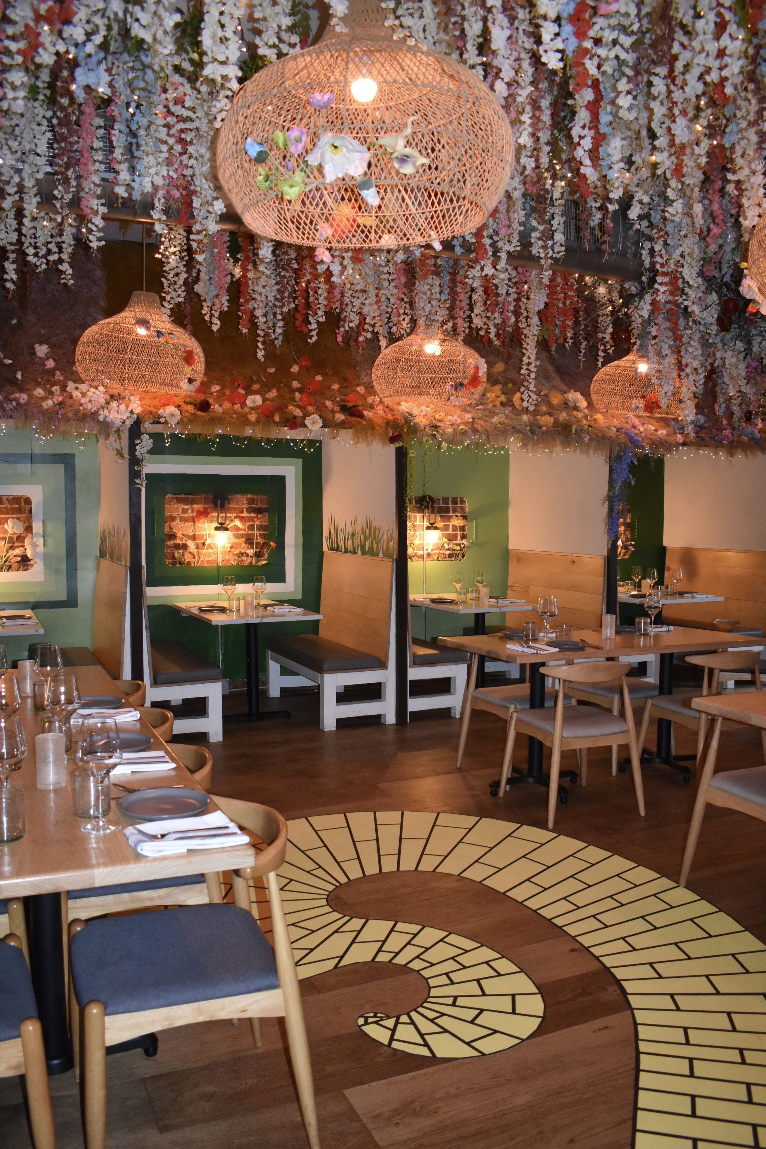 A restaurant interior with hanging wicker light fixtures, floral decorations hanging from the ceiling, and a patterned tile floor. Wizard of Oz themed decorations with yellow brick road, munchkinland huts, and overgrown florals.