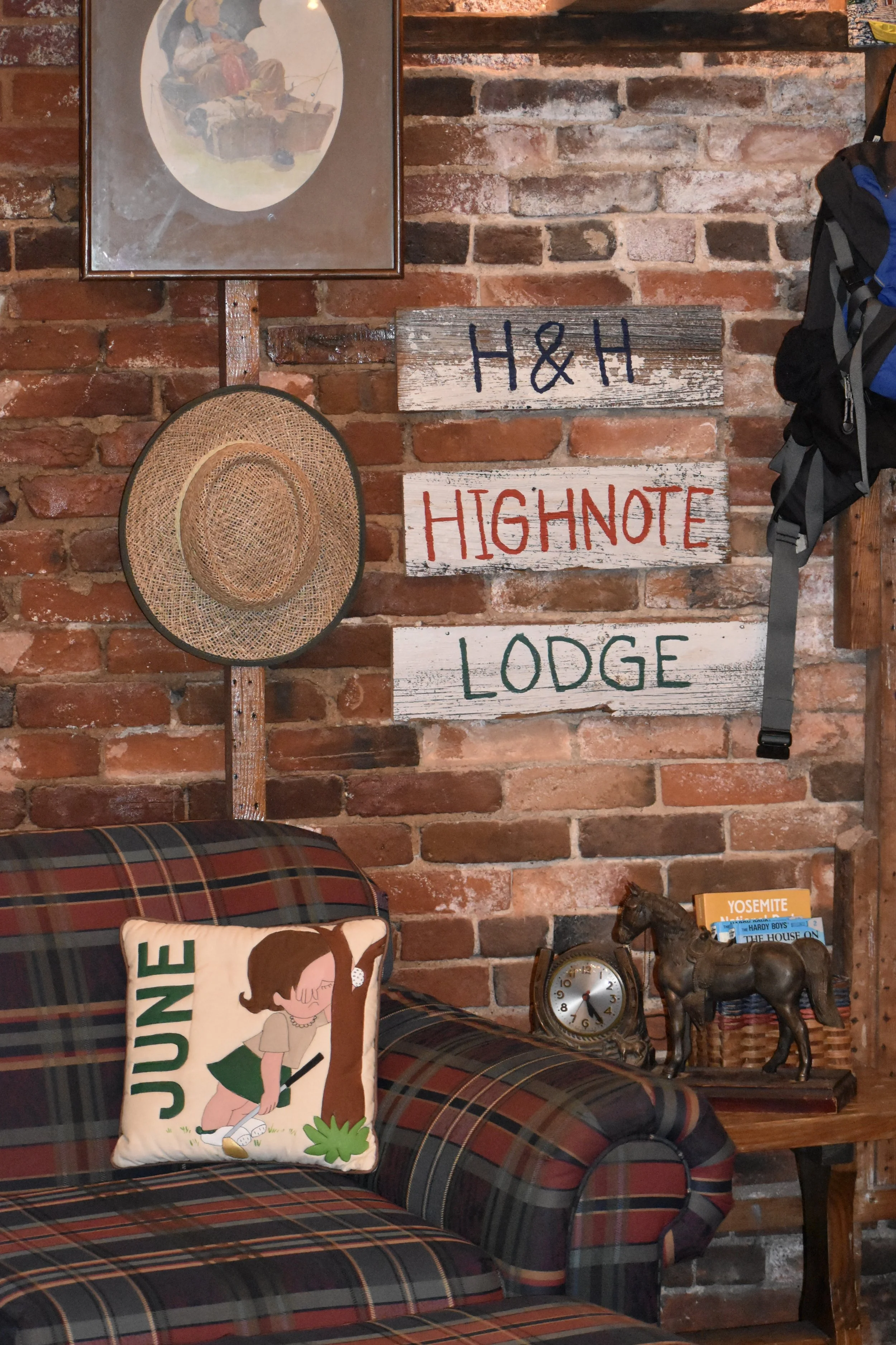 Decorative interior scene featuring a brick wall with signs reading 'H&H Highnote Lodge,' a straw hat, a framed painting, a plaid sofa with a "June" decorative pillow, a vintage clock, a small horse figurine, a stack of books, and a backpack hanging 