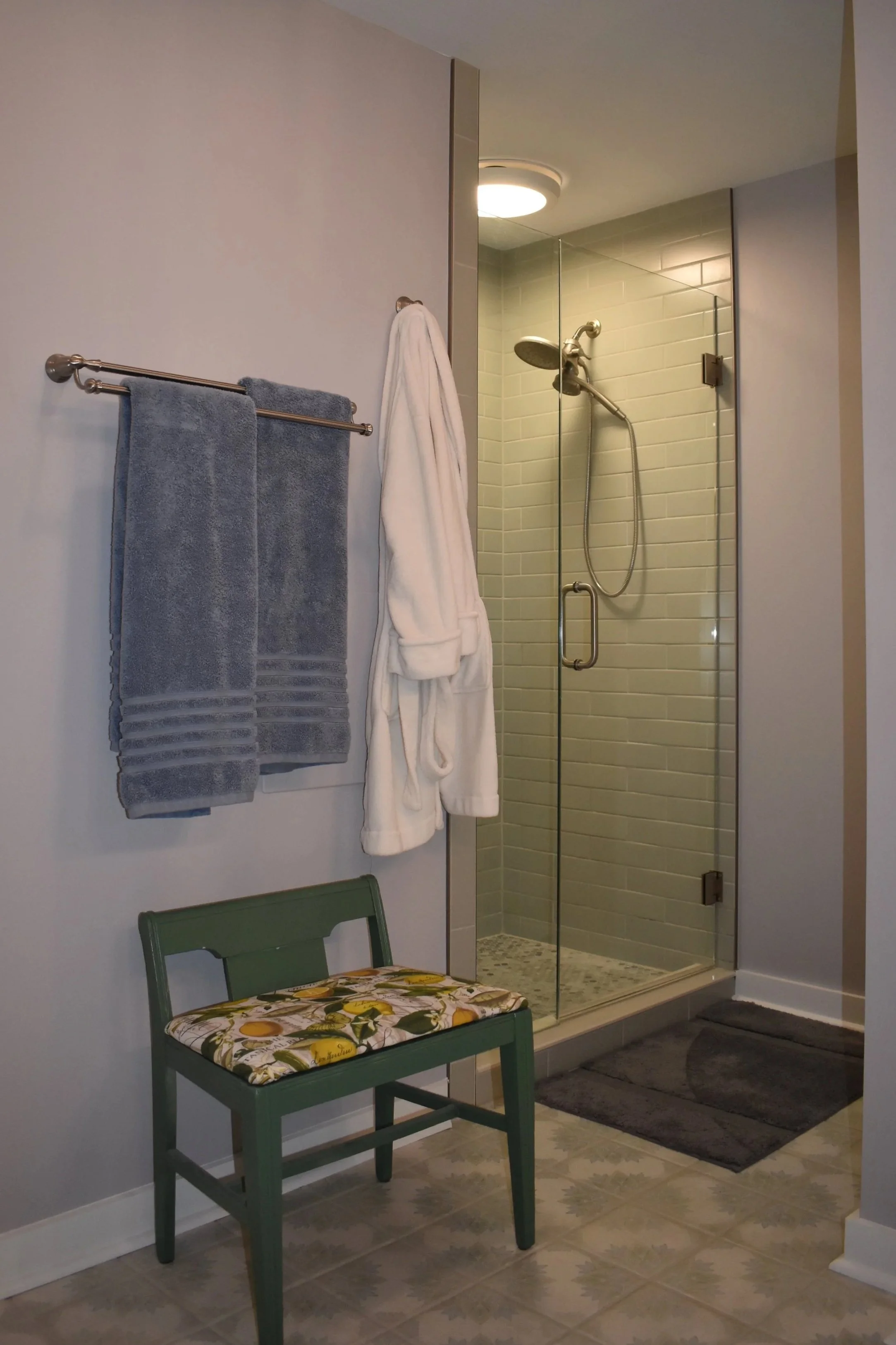 A bathroom with a walk-in shower, towels hanging on a wall, a white bathrobe, a green bench with a floral cushion, and a gray rug on tile flooring. Marietta GA bathroom remodel.
