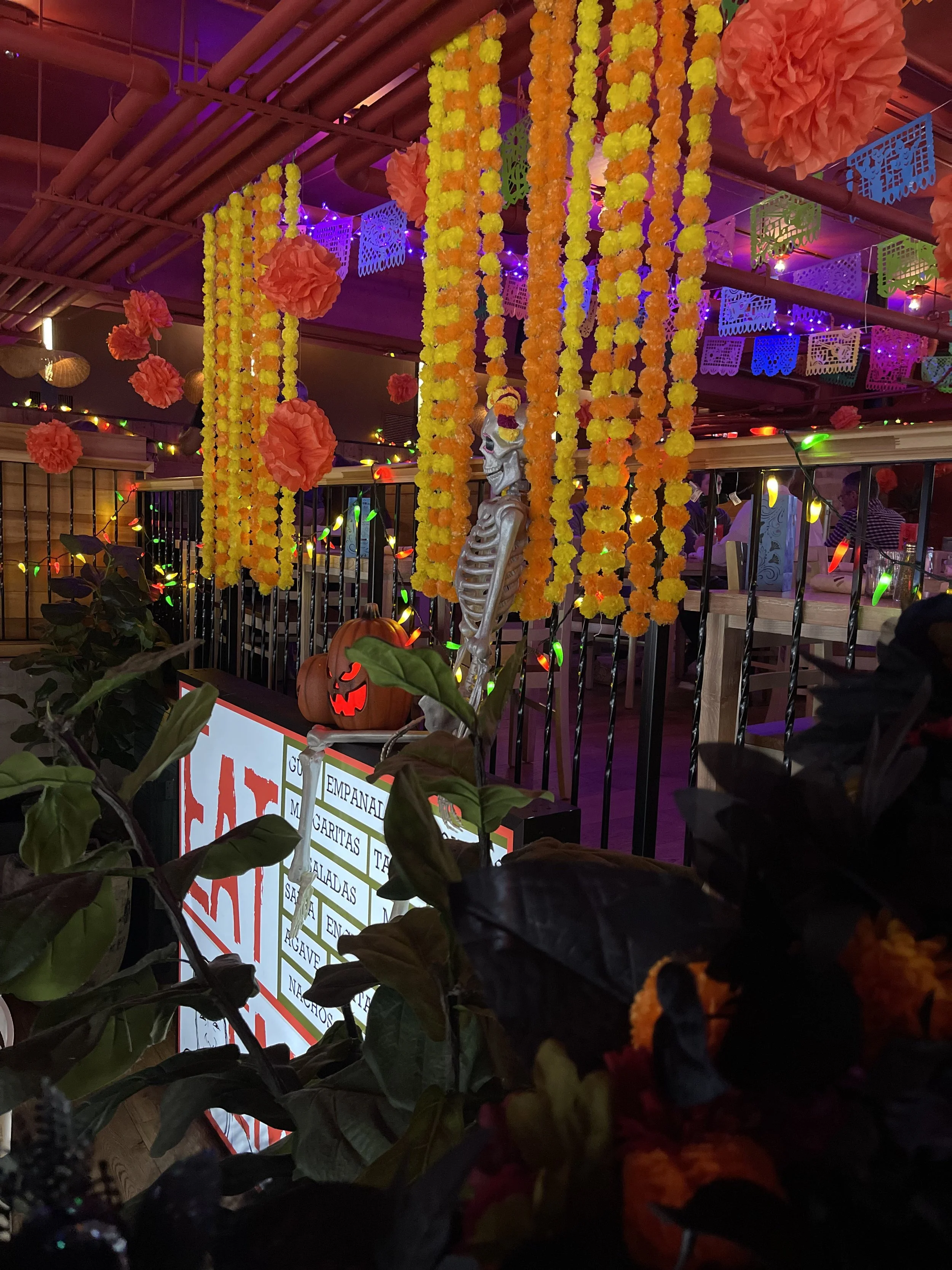 Halloween party decorations with orange and yellow flower garlands, pink tissue paper flowers, a skeleton figure, a carved pumpkin with a carved face, and festive lighting in a decorated indoor space.
