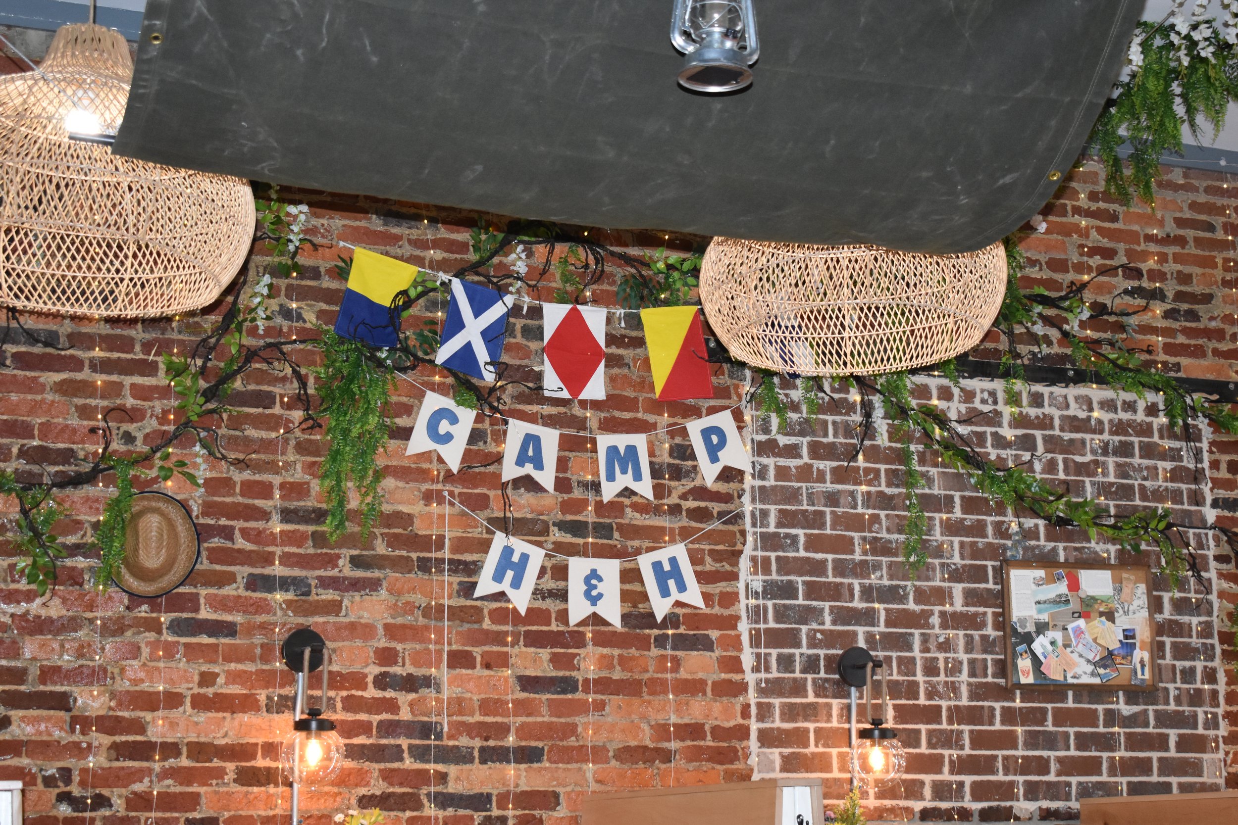 Decorative indoor space with a brick wall, hanging wicker lamps, green foliage, string lights, and a banner that reads 'Camp Hёh' with nautical and camping flags. Hamp & Harry's Marietta Square.
