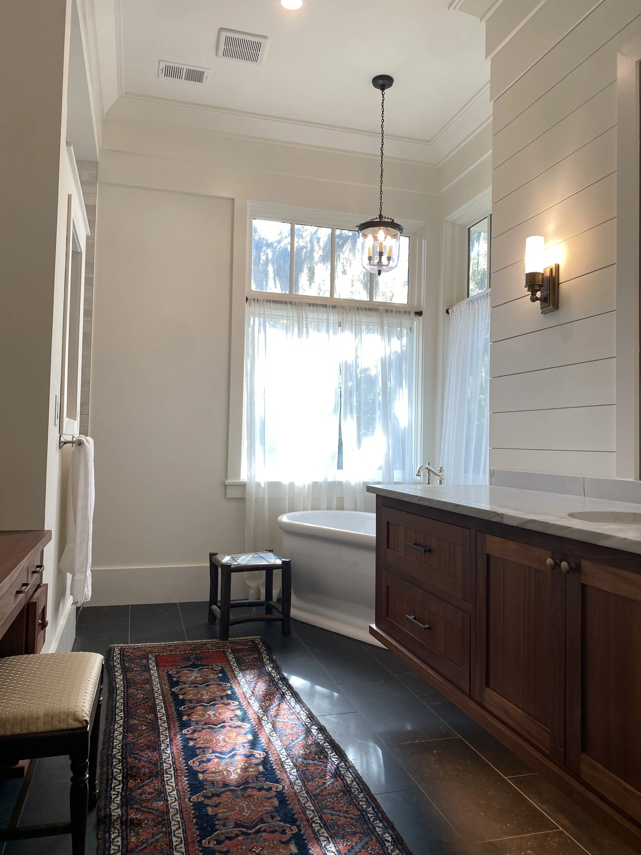 Bathroom with a bathtub near a large window, wooden vanity with a marble countertop, vintage rug on dark tile floor, hanging light fixture, and wall sconces. Hilton Head Island Interior Design.