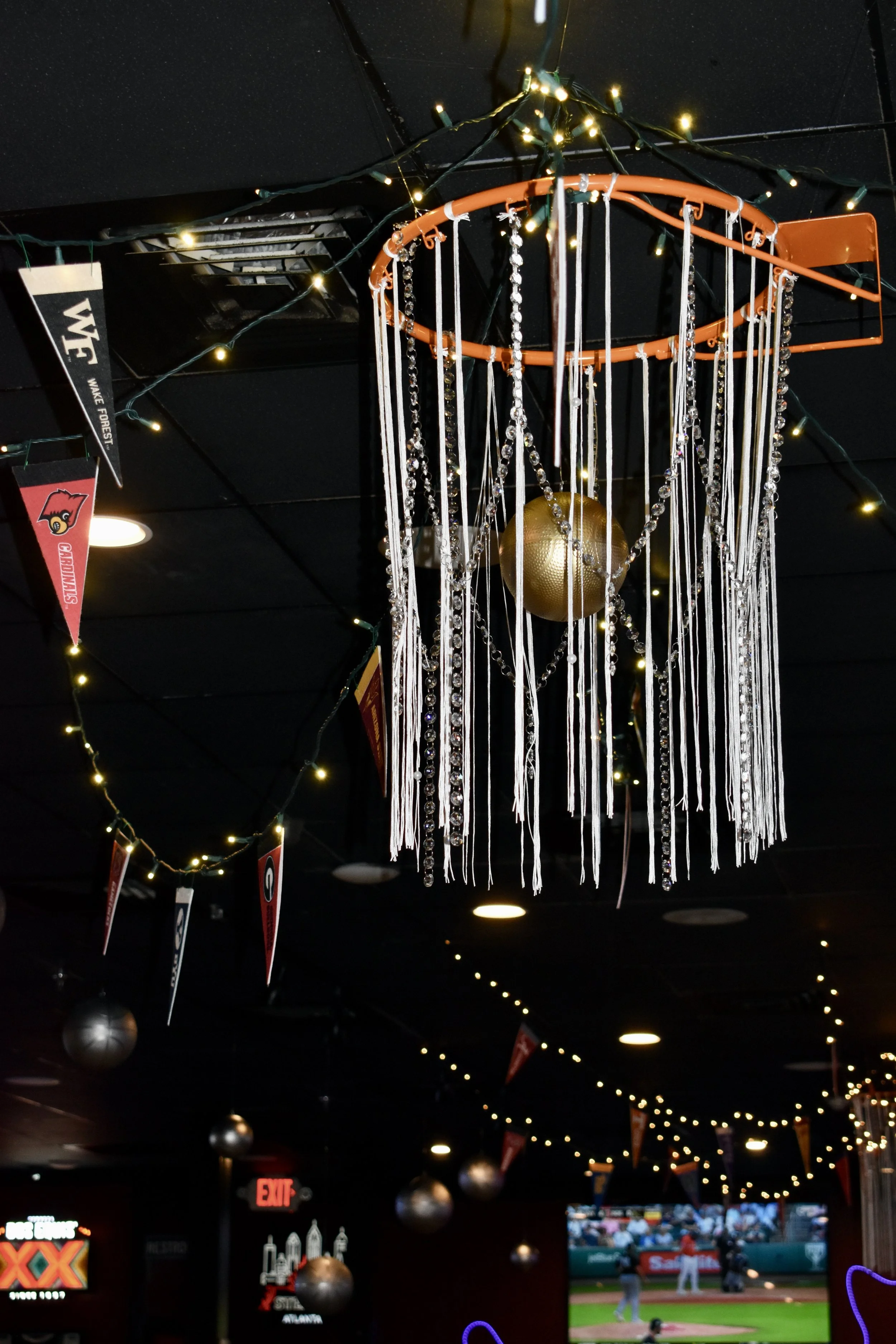 Decorative chandelier in sports bar with string lights and University of Louisville banners, with a television showing a baseball game in the background. March Madness