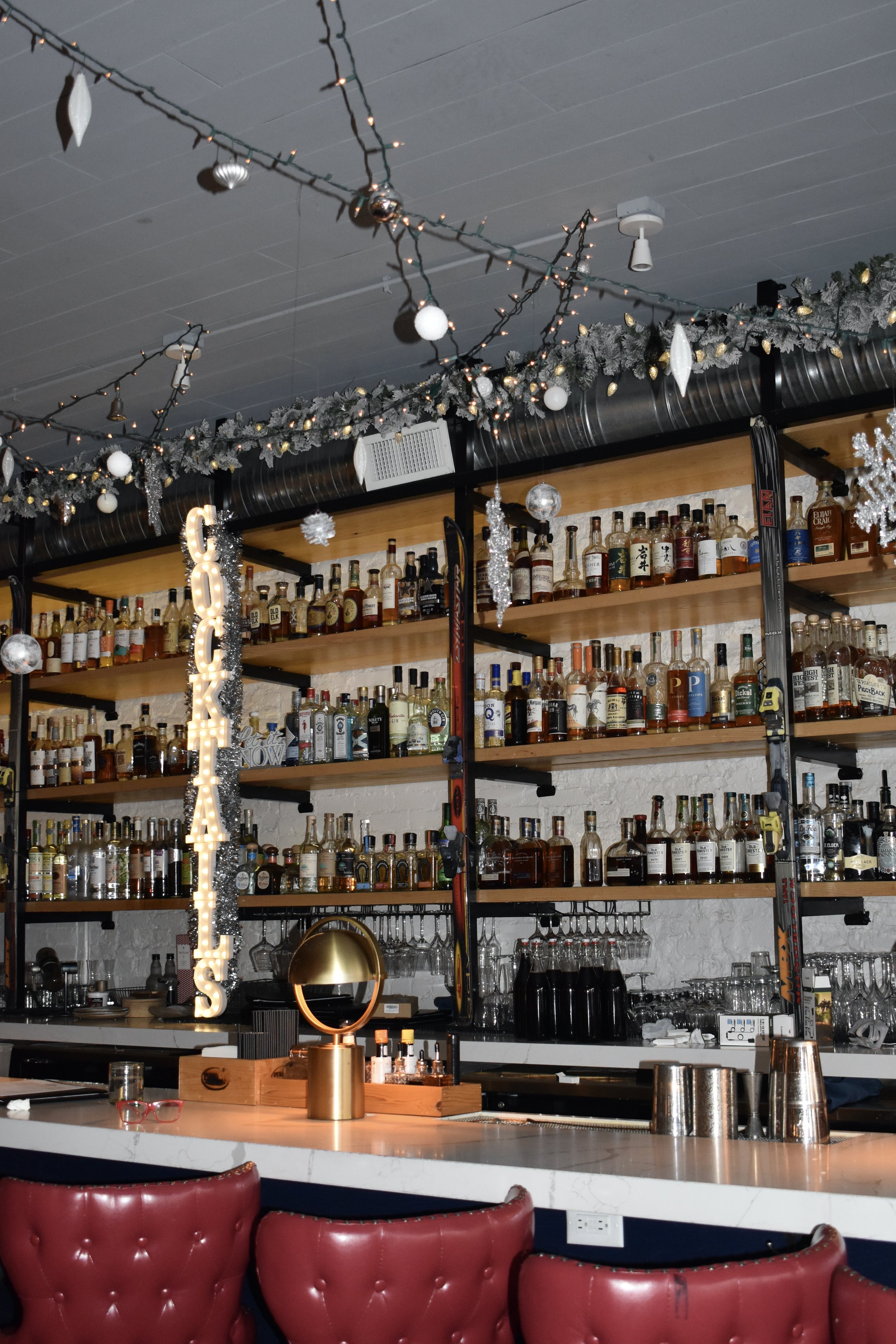 Bar area decorated with Christmas ornaments, including hanging ornaments and garland, and a lit 'Look at This' sign with lights. Bottles of liquor are displayed on shelves behind the bar, and red cushioned bar stools are in the foreground.