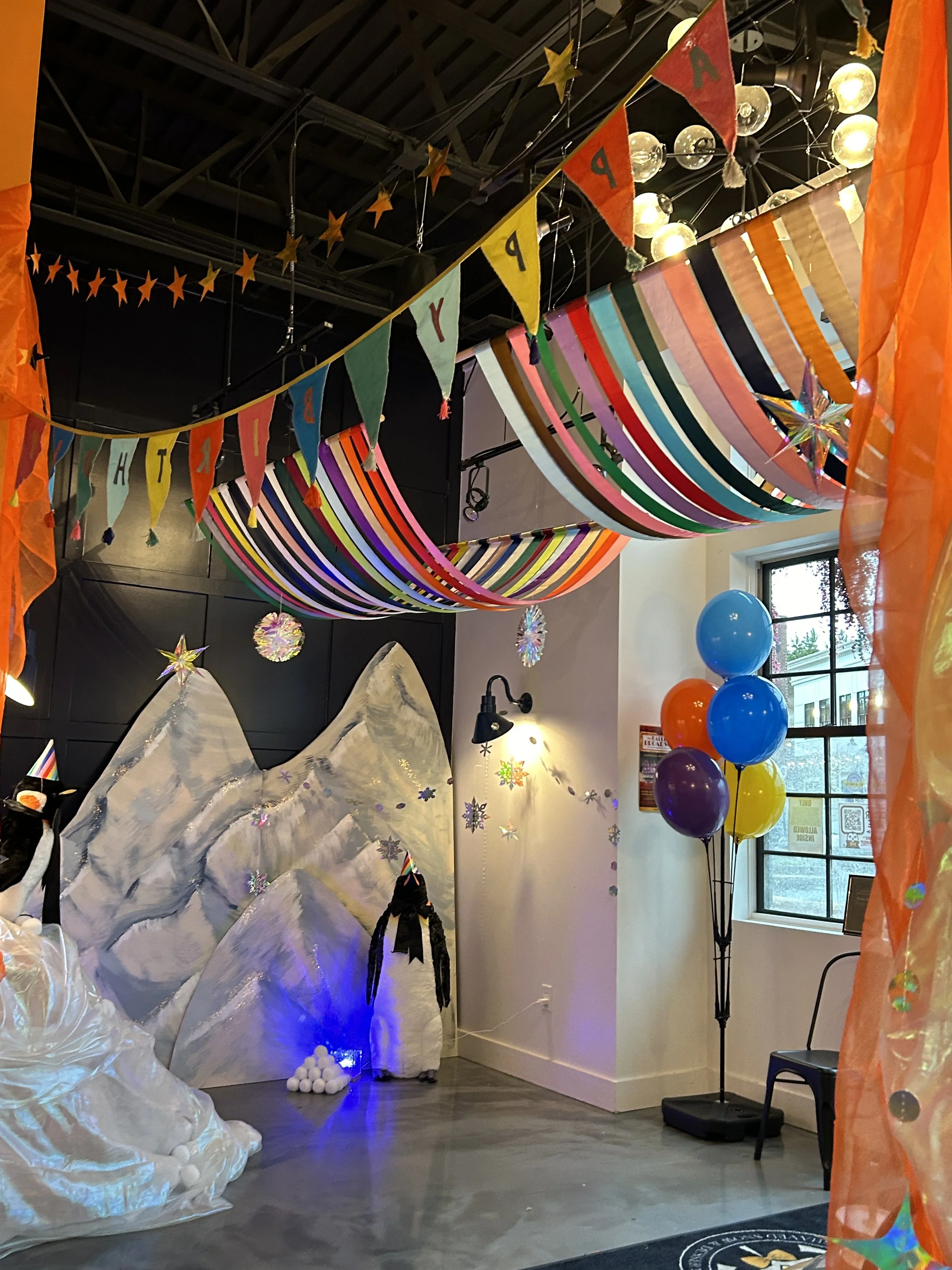 Colorful party decorations with rainbow-colored fabric hangings, banners spelling out 'HAPPY PARTY,' balloons, snow-capped mountain backdrop, and paper snowflakes, creating a festive indoor scene.