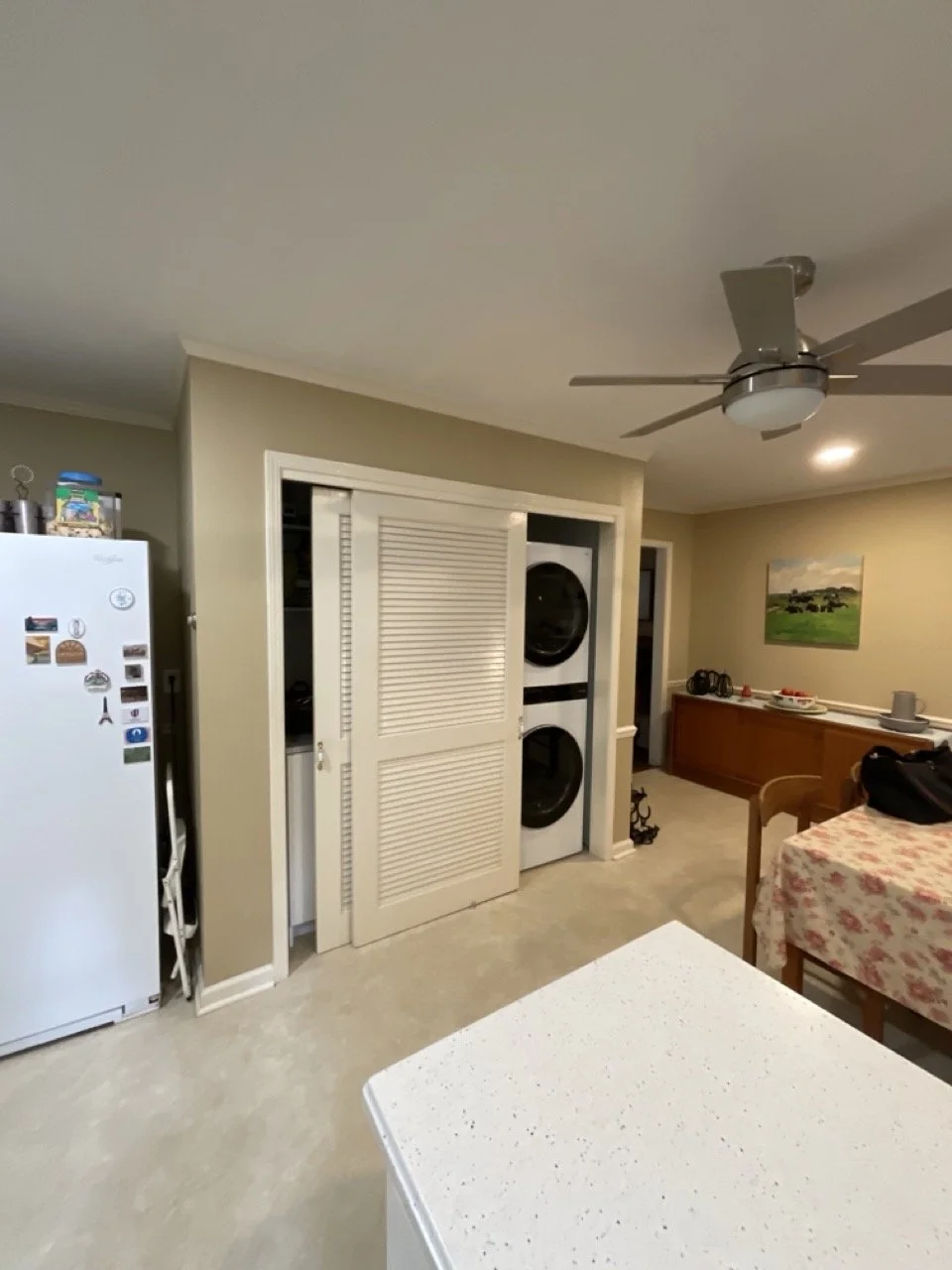 Kitchen with a white refrigerator, a stacked washer and dryer behind a closet door, a ceiling fan, a dining table with a floral tablecloth, and a painting on the wall.
