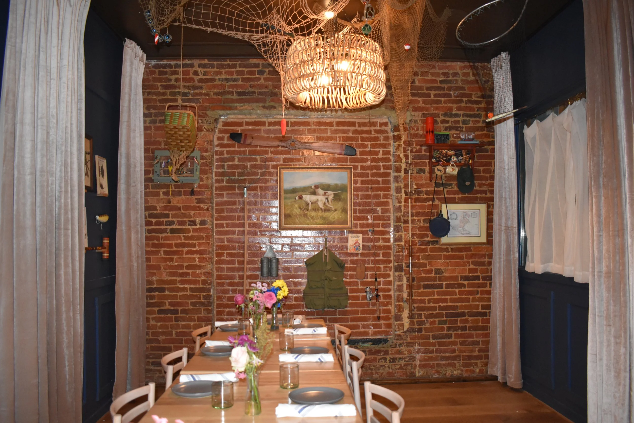 Interior of a rustic dining room with a long wooden table set with plates, glasses, and a flower centerpiece, surrounded by white chairs. The room features a brick wall decorated with vintage and outdoor-themed items such as a painting of dogs, a kay